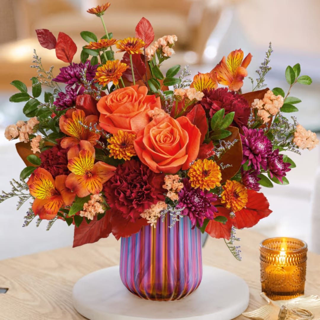 Orange roses and mixed fall flowers in a striped glass vase