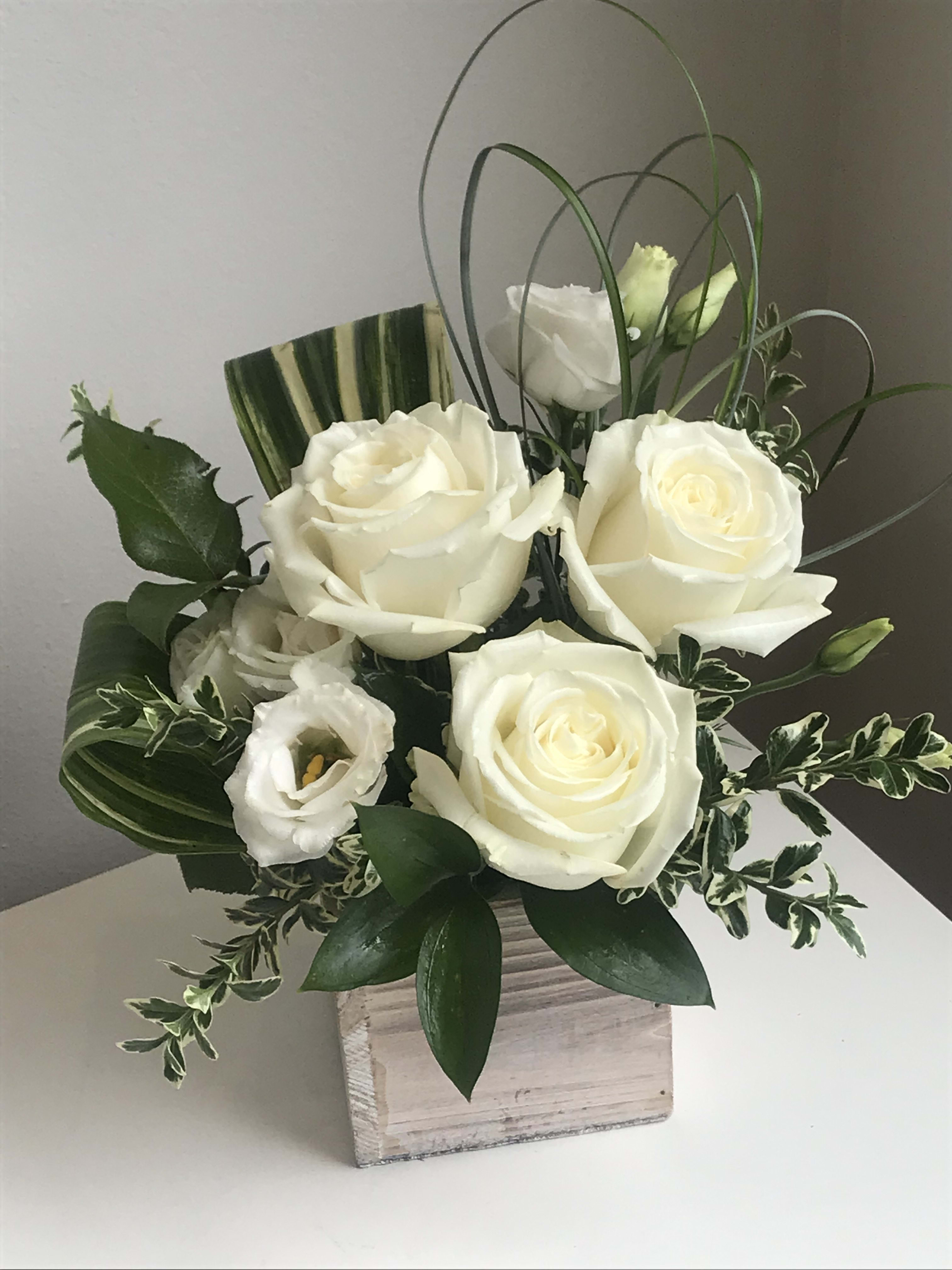 White roses and lisianthus in a square wooden vase with looping grass accents