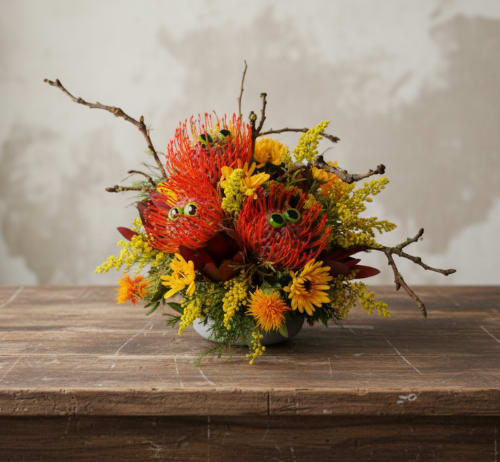 Bright orange and yellow floral arrangement with branch accents in a low bowl