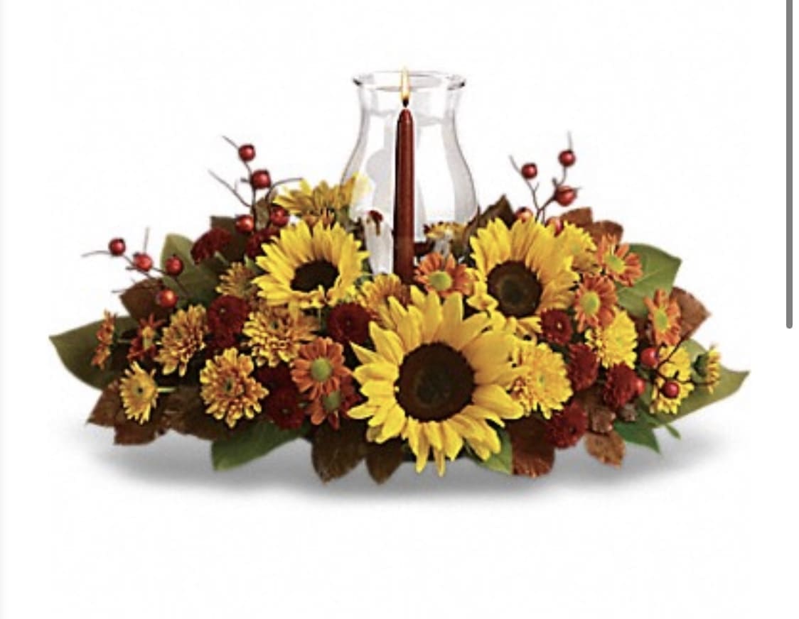 SUNFLOWER CENTERPIECE - This centerpiece maximizes the joy of sunflowers by arranging them with a beautiful mix of fall flowers, foliage and a cranberry hued candle under a hurricane glass. sunflowers,spray roses, broze and rust pompoms, red berries,lemon leaf and a tapered candle inside a hurricane vase.