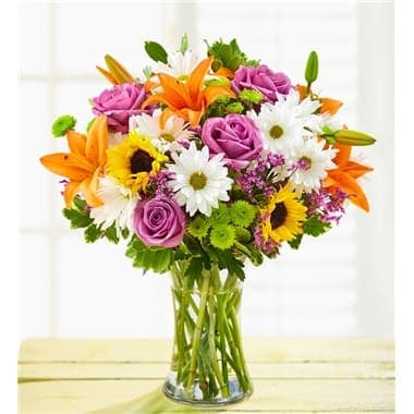 Mixed bouquet of roses, lilies, daisies, and sunflowers in a glass vase