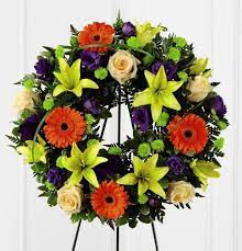 Circular floral wreath with orange gerberas, yellow lilies, cream roses, and purple blooms