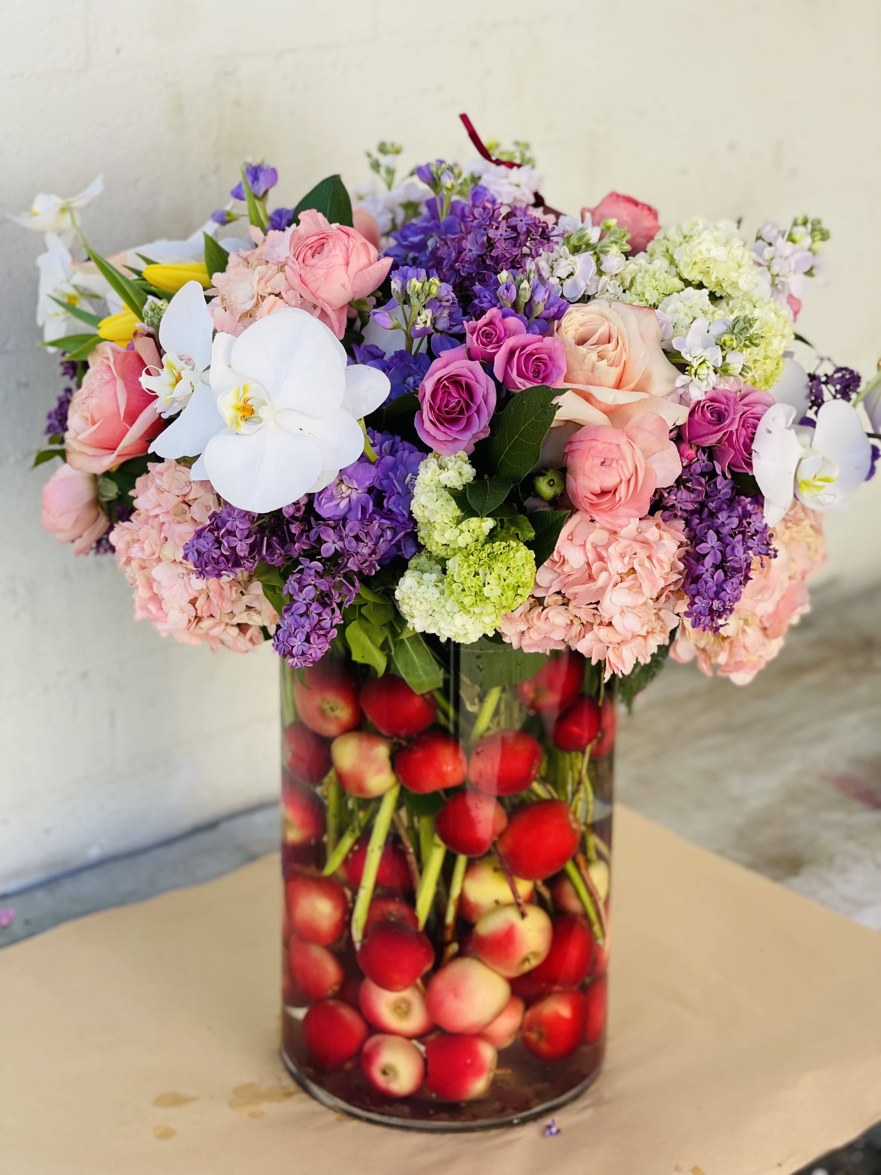 Mixed pink, purple, and white flowers in a tall glass vase filled with apples