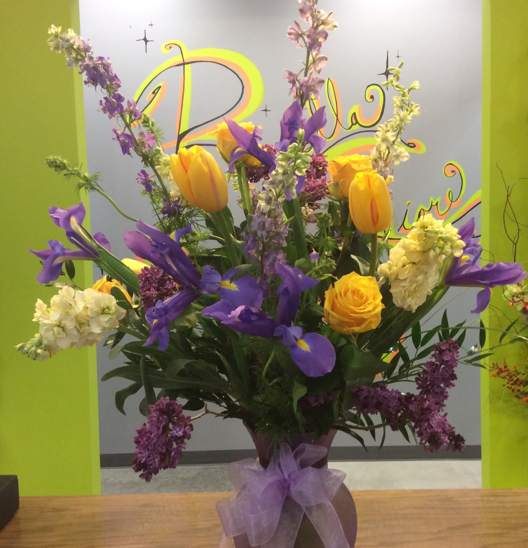 Tall bouquet of yellow tulips, purple irises, and lilac in a vase with a purple bow
