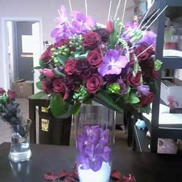 Purple and burgundy floral arrangement in a clear vase with a purple orchid cylinder centerpiece