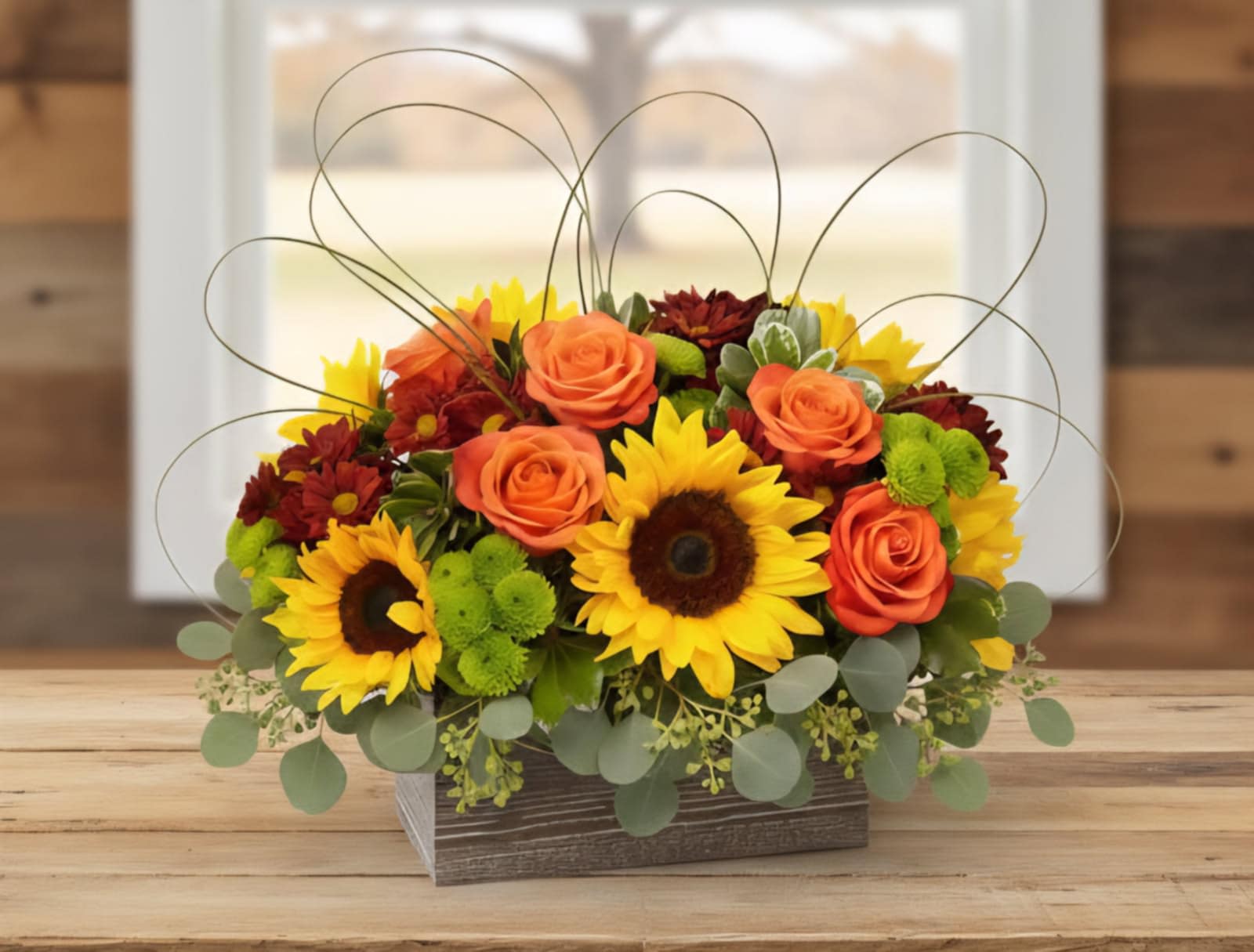 Low arrangement of sunflowers, orange roses, and mums in a rustic wooden box with looping wire accents.