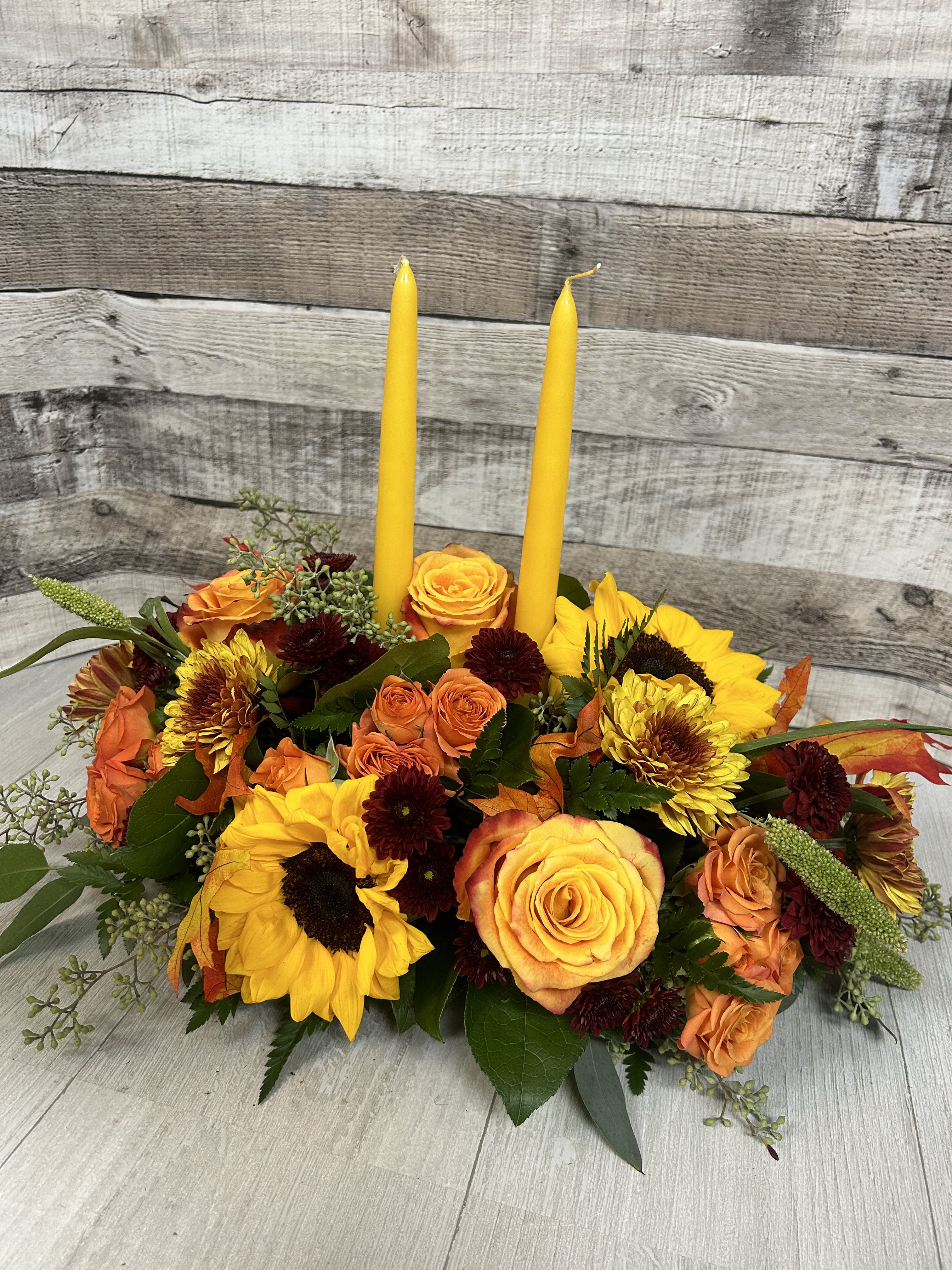 Glowing Gathering - This centerpeice brings a variety of florals like sunflowers, orange spray roses and vibrant mums. This assortment brings a cozy vibe wherever it is placed.