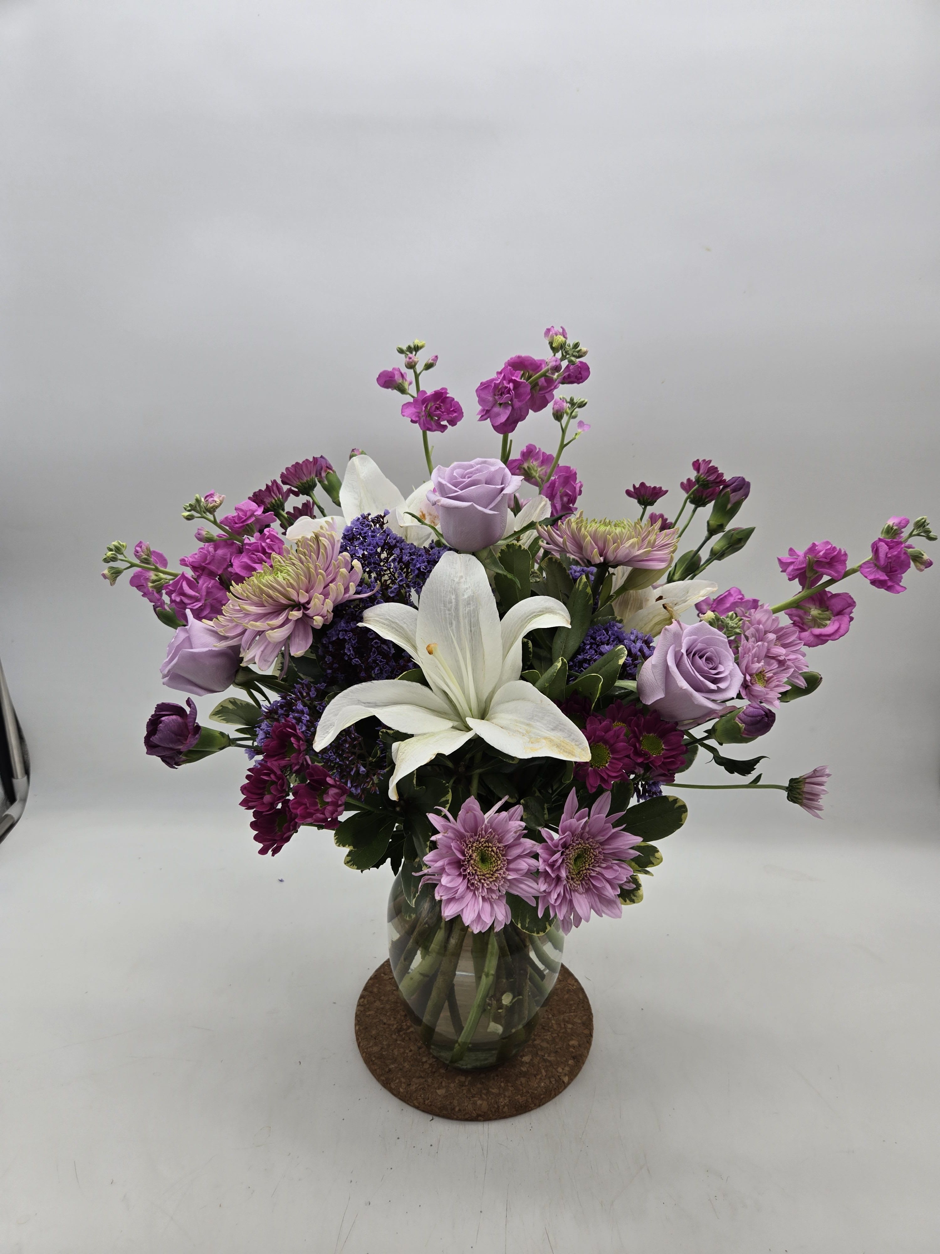 The Dreamscape - Soft lavender roses and airy daisy-like blooms tumble around two prominent white lilies to create a calm, romantic flower arrangement perfect for a birthday bouquet, anniversary flowers, or a thoughtful just-because gift. The Dreamscape's gentle purple, pink and white palette reads both fresh and serene, making the recipient feel truly seen-comforted, celebrated, or quietly surprised by a moment that feels carefully curated.  Arranged in a clear glass vase with visible stems and loose greenery that spills slightly around the edges, this rounded bouquet has a natural, garden-style shape with layered texture. Lilies provide a graceful focal point while the lavender roses and daisy-shaped accents add softness and delicate contrast; touches of green keep the look fresh and airy. Sized for everyday gifting, it's an elegant choice for spring celebrations, thank-you gestures, or sympathy arrangements-an easy-to-care-for display that brightens a bedside table, entryway, or office desk.