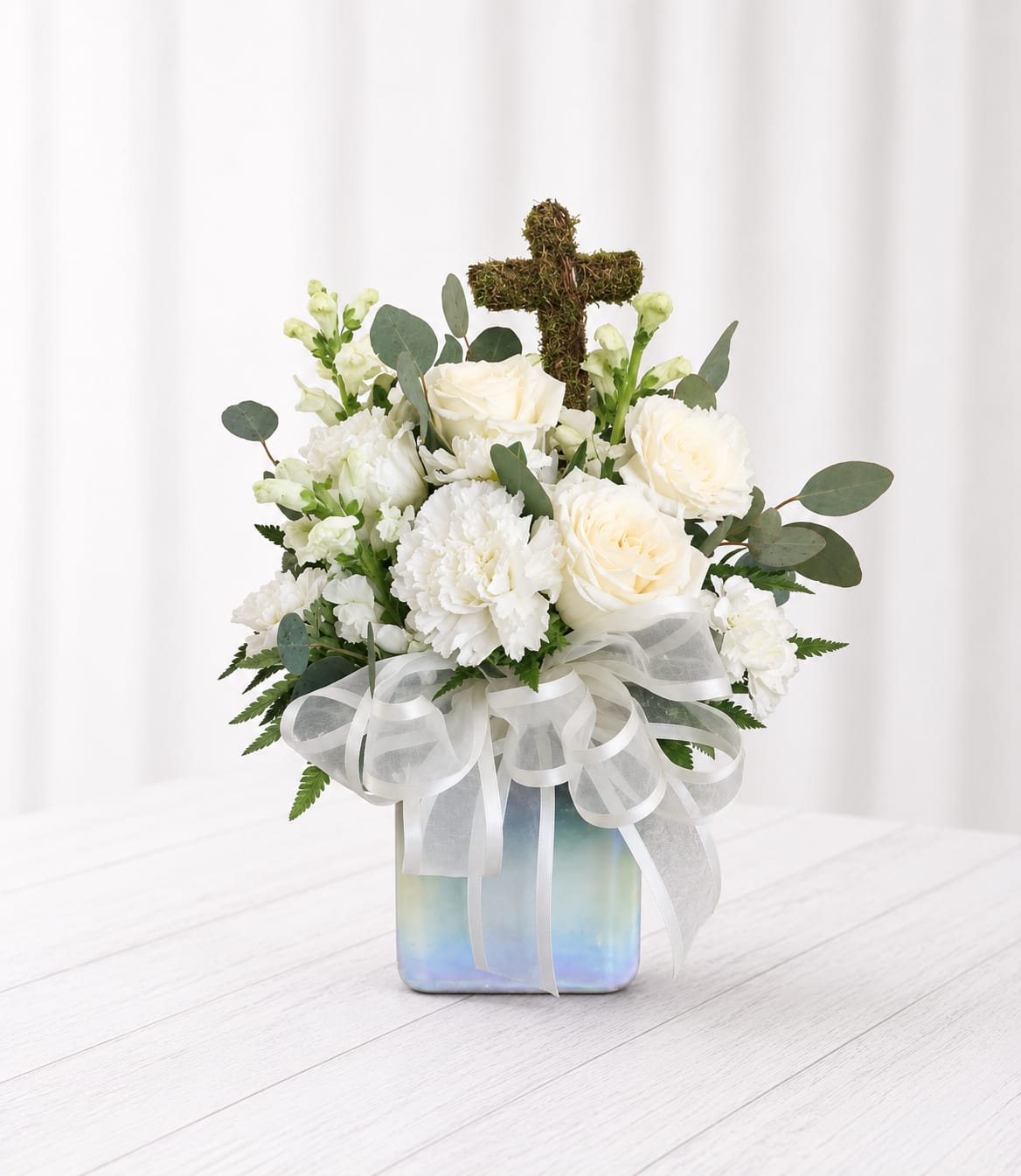 Faith - A serene, compact sympathy arrangement centered on soft white roses and carnations, Faith conveys quiet comfort for a funeral, memorial service, or any moment you need to express condolence. The upright design is accented with pale green snapdragon spikes and eucalyptus, and a moss-covered cross (currently substituting a ceramic cross) rising from the back gives this piece a gentle religious touch-appropriate for Memorial Day remembrances or other faith-based services. 