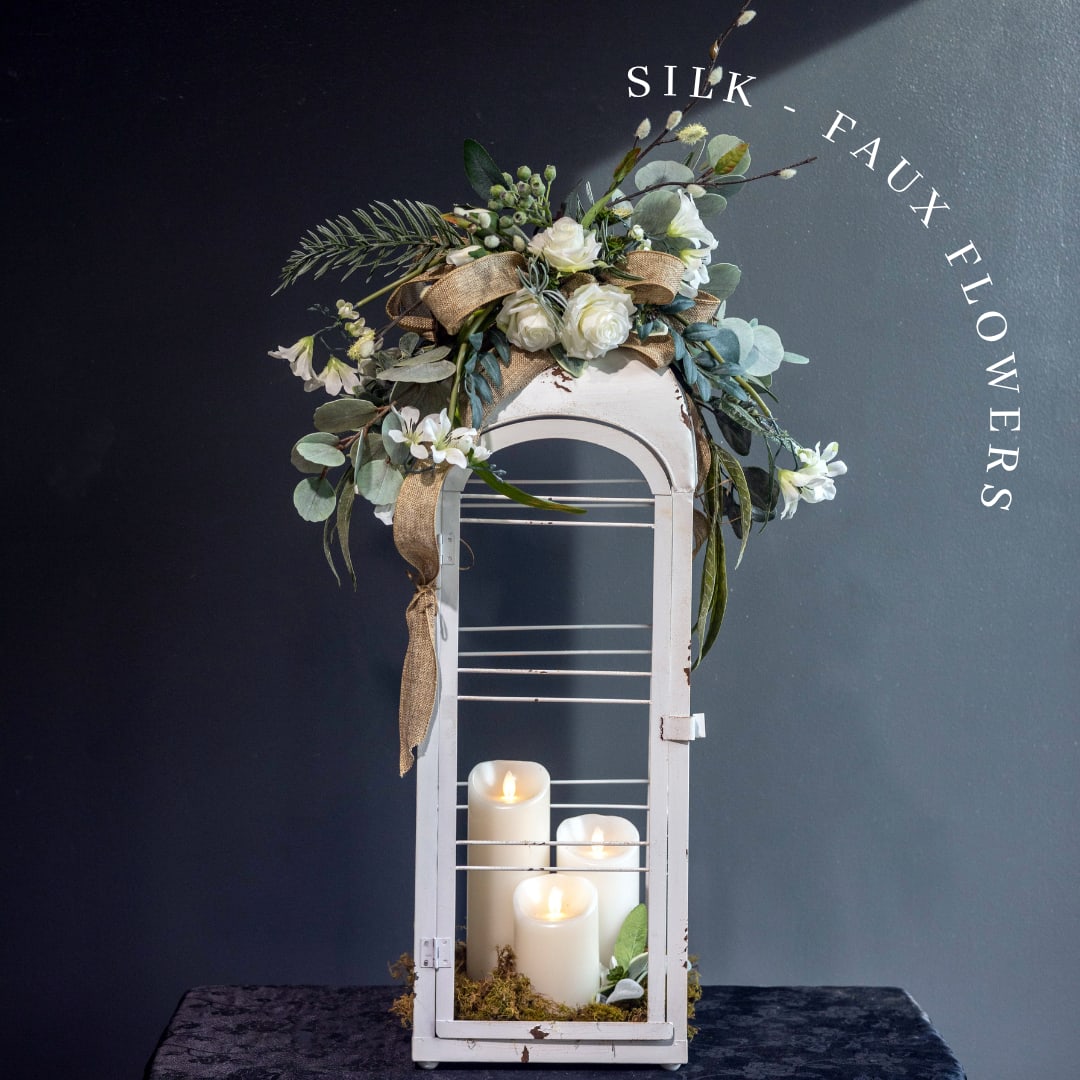 A Memories Glow: Silk - Faux Flowers - Honor a cherished life with A Memory's Glow, a timeless faux sympathy lantern designed to bring warmth, peace, and lasting remembrance. This elegant keepsake features a distressed white lantern adorned with premium silk white roses, eucalyptus, delicate greenery, and a rustic burlap ribbon, creating a graceful tribute that complements any memorial or celebration of life.  Crafted with lifelike florals that require no maintenance, this arrangement offers a beautiful, lasting expression of sympathy.