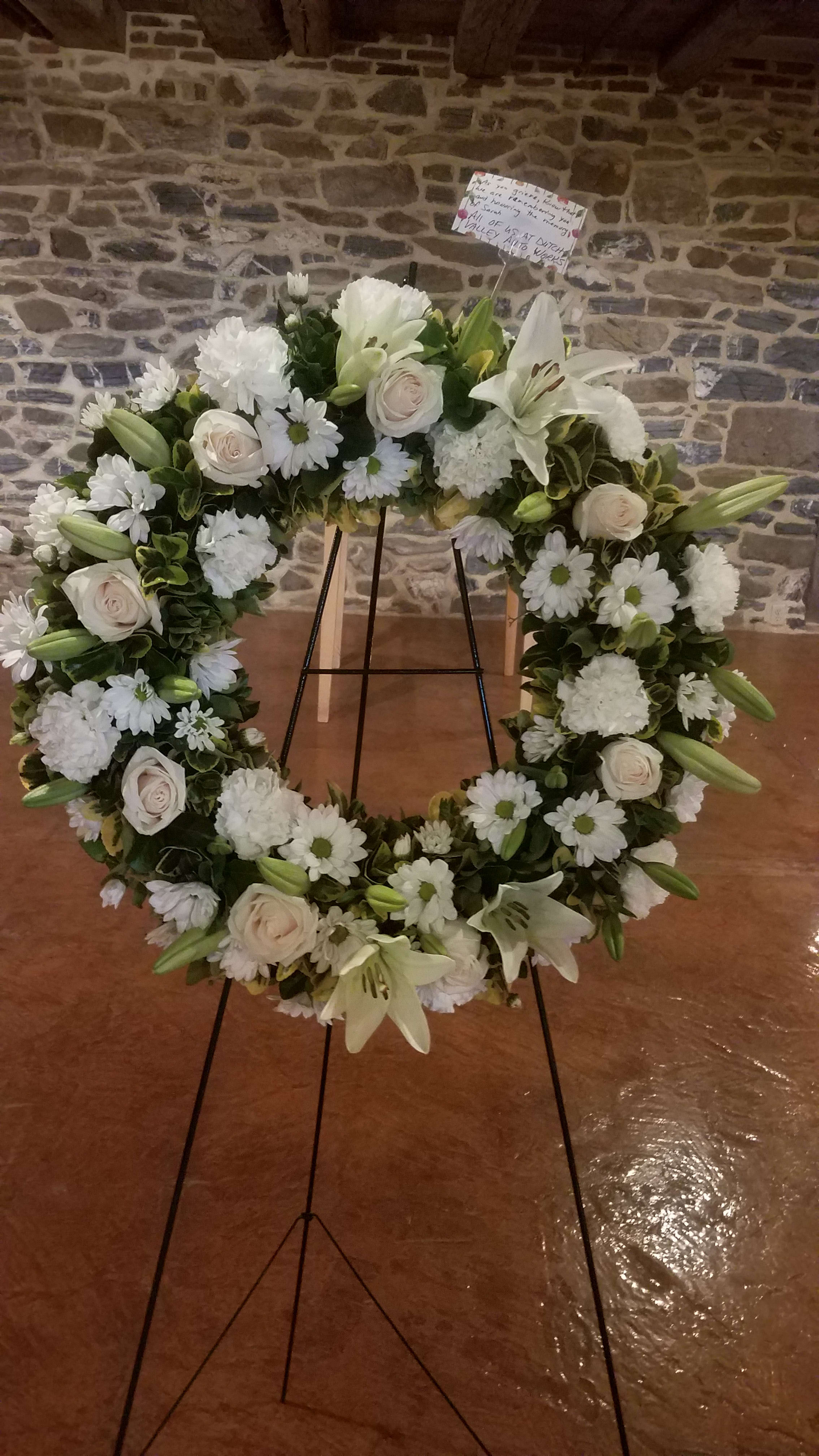 White floral wreath on a stand with lilies, roses, and daisies