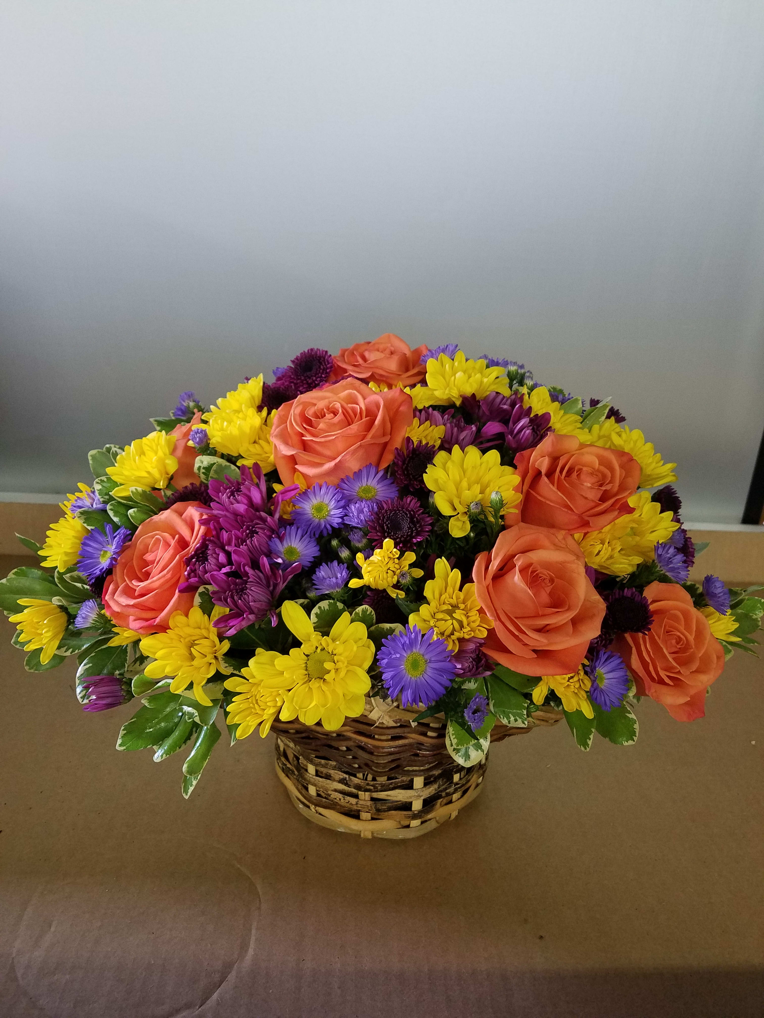 Basket arrangement of orange roses, yellow daisies, and purple flowers