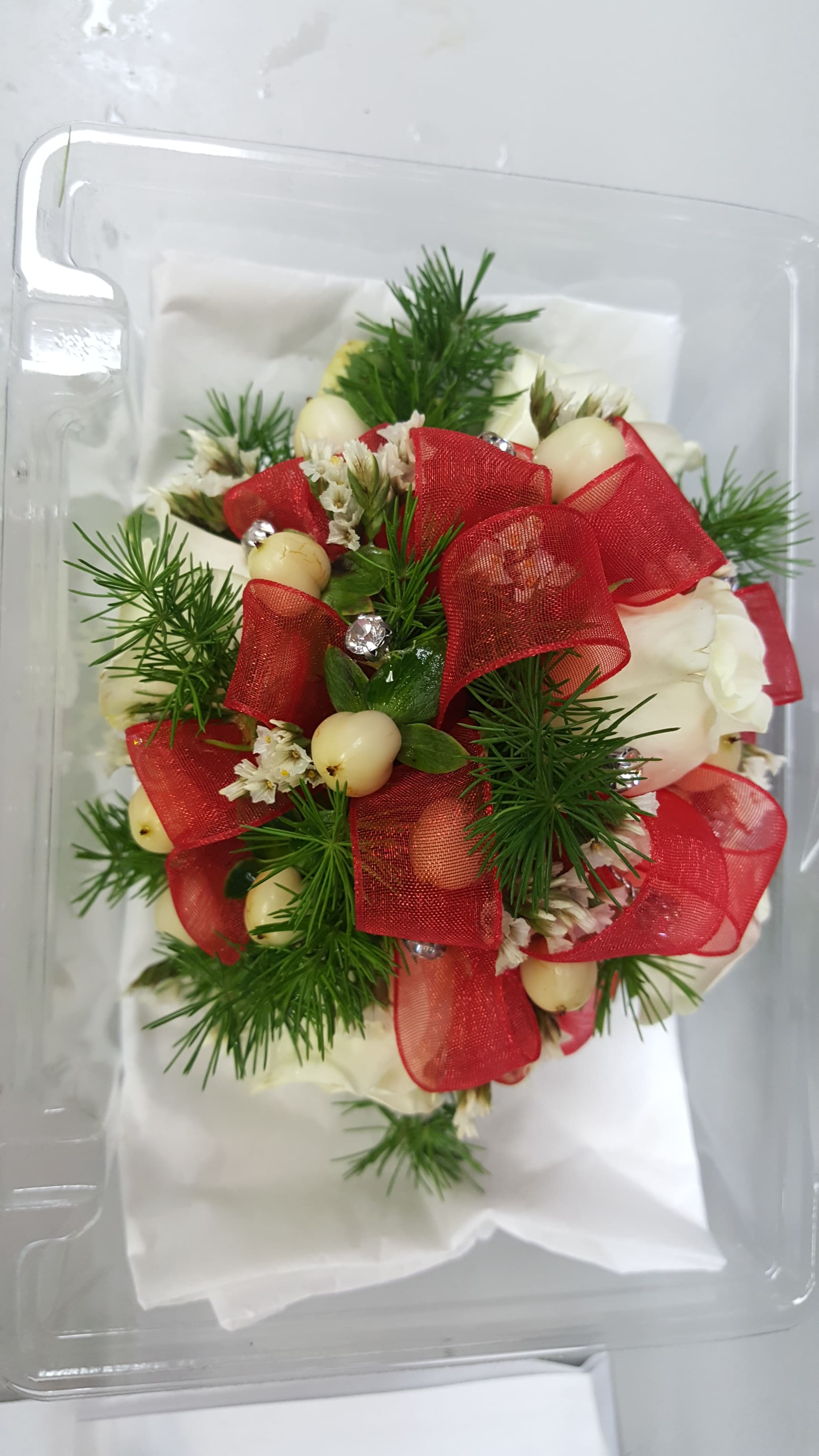 Small white floral arrangement with red ribbon and berries in a clear plastic box