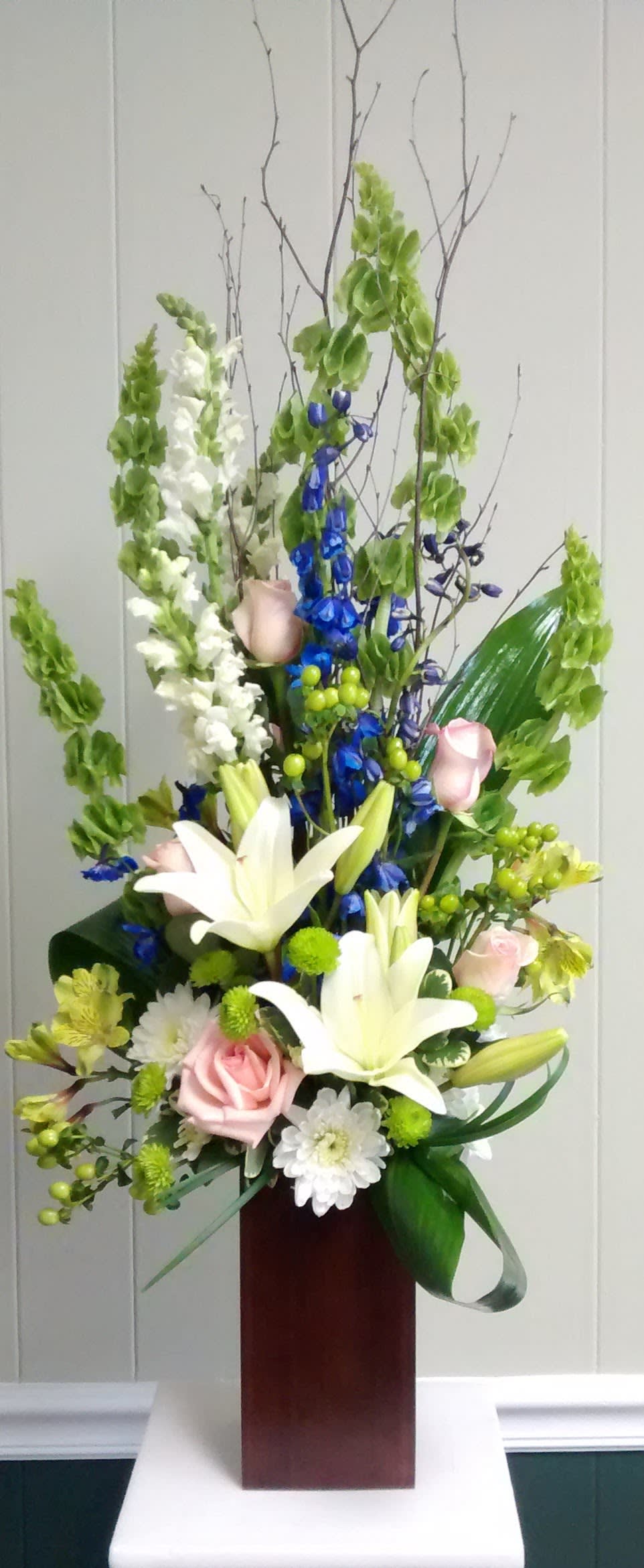 Tall floral arrangement with white lilies, pink roses, and blue flowers in a wooden vase