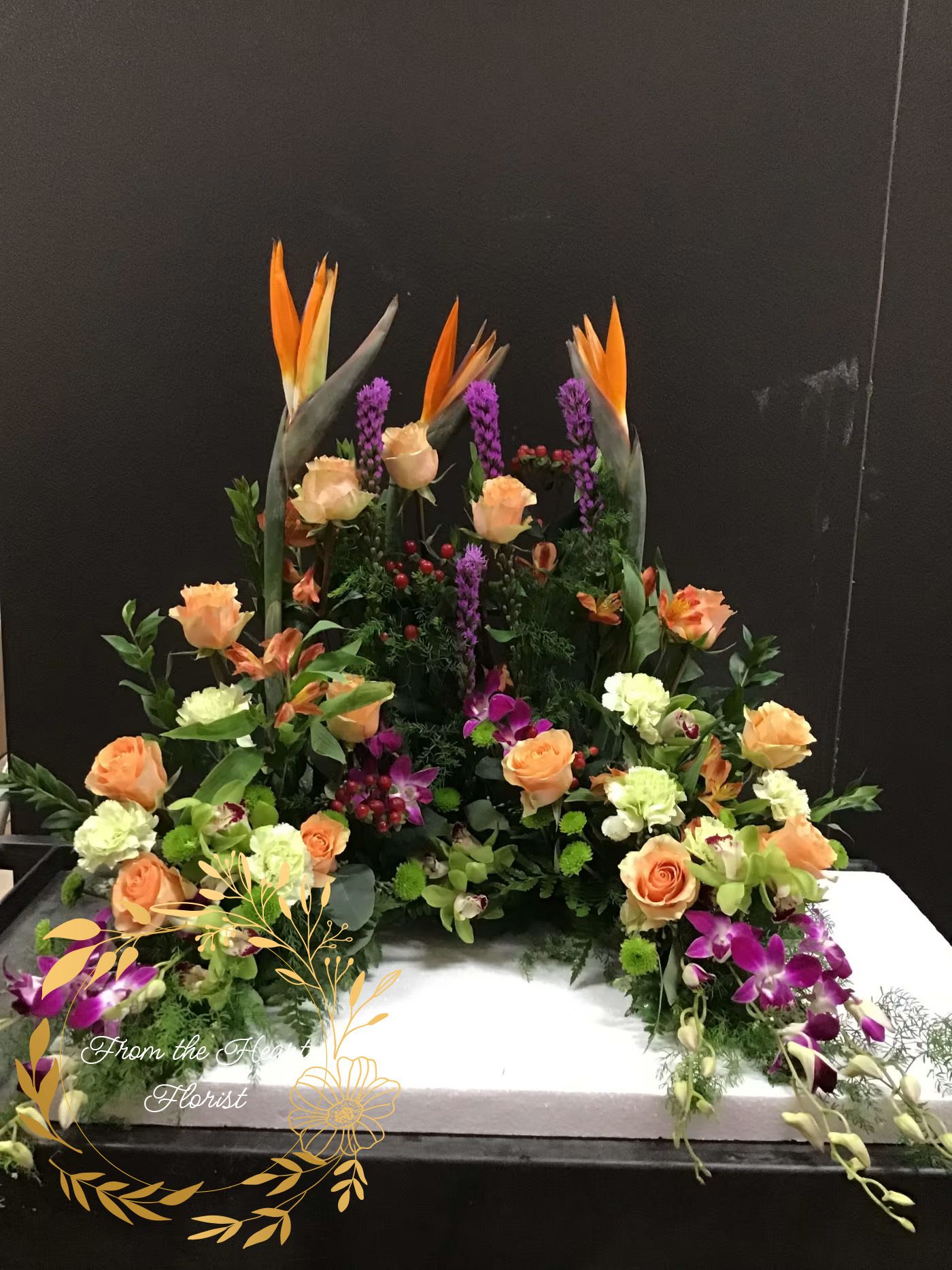 Large floral arrangement with orange roses and purple orchids on a white base