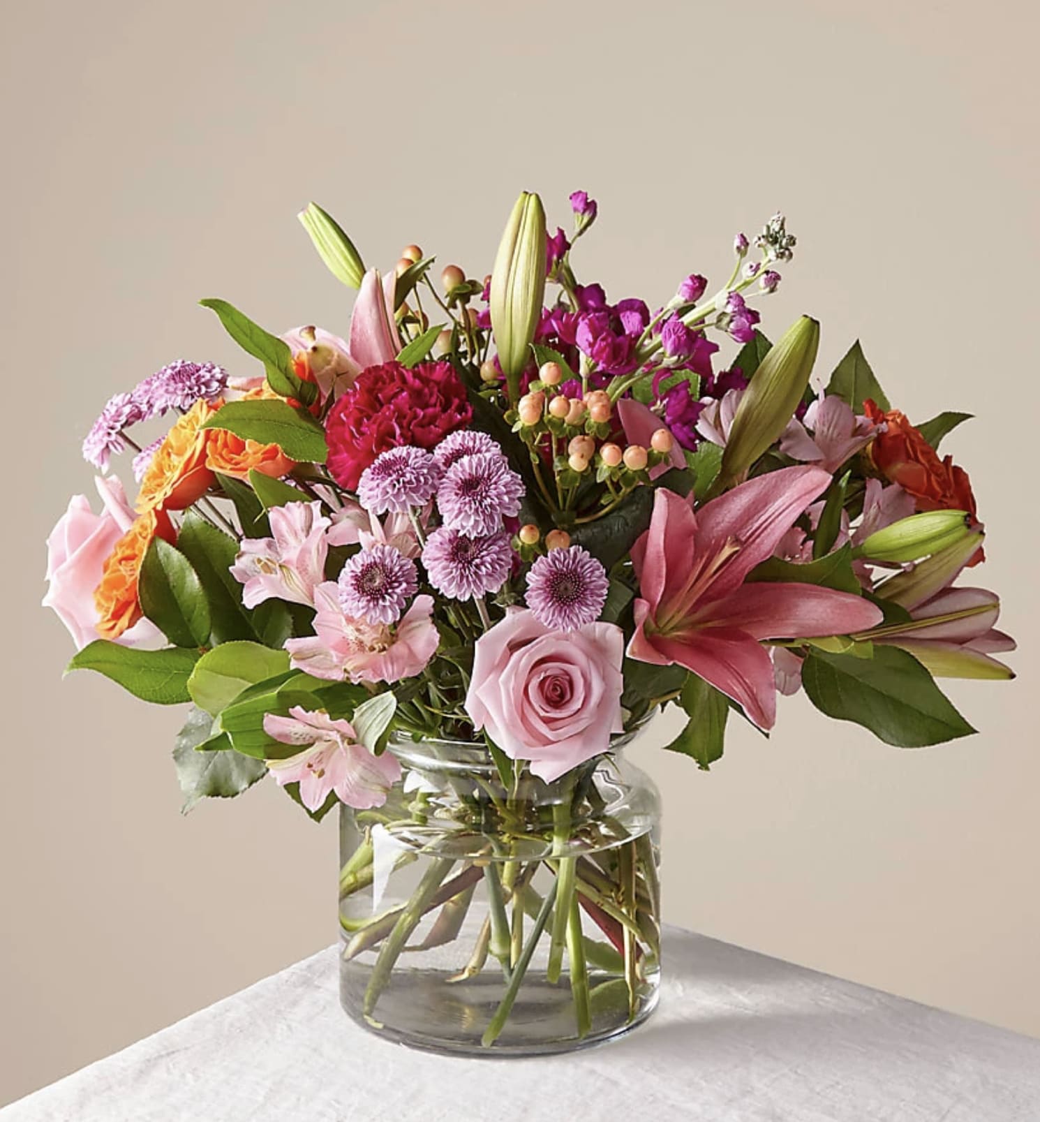 Mixed pink, purple, and orange flower arrangement with roses and lilies in a clear glass vase