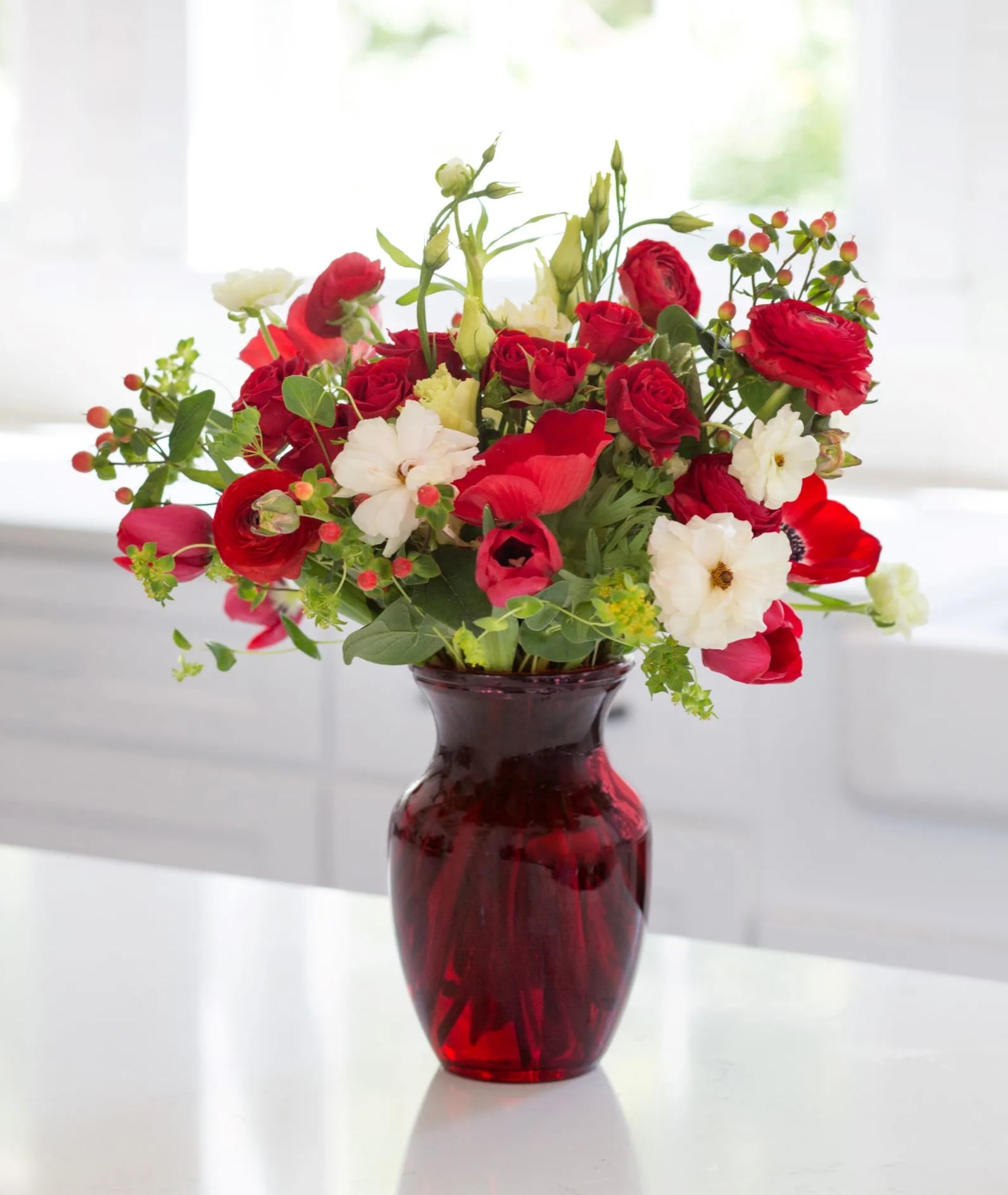 Red and white mixed flower arrangement with berries in a red glass vase on a white countertop