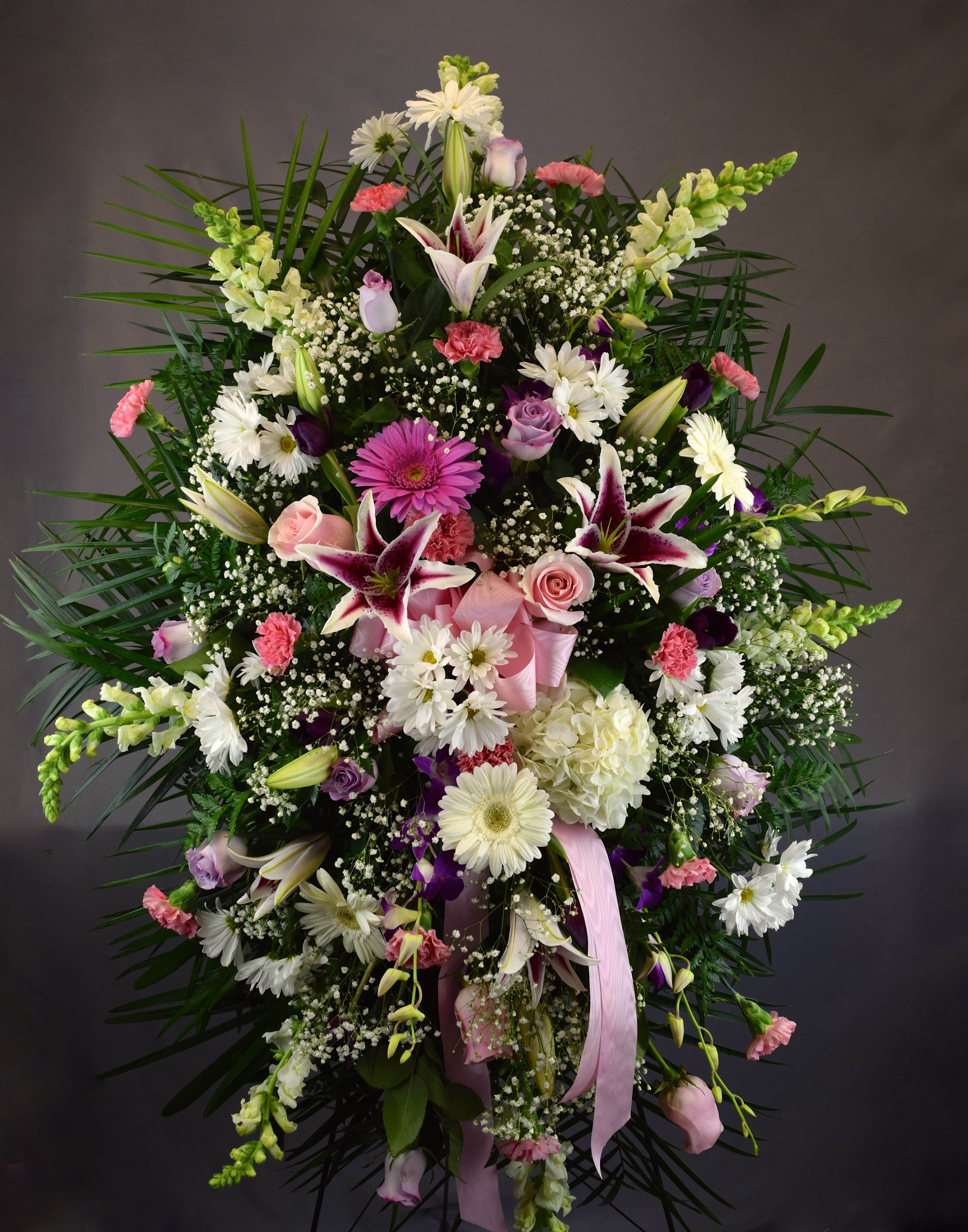 Large standing floral spray with lilies, daisies, roses, and pink ribbon