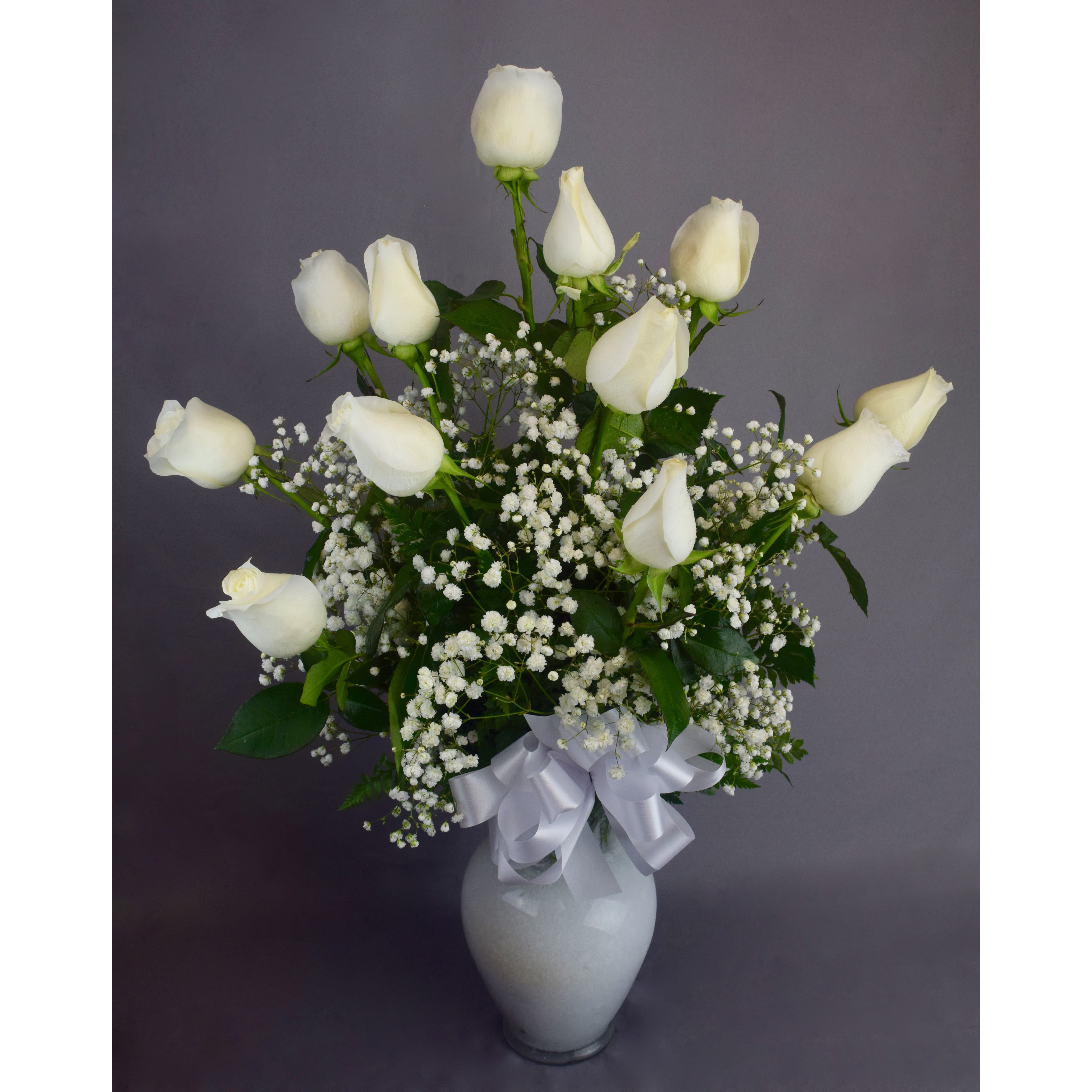 White roses arranged in a white vase with baby's breath and a white ribbon