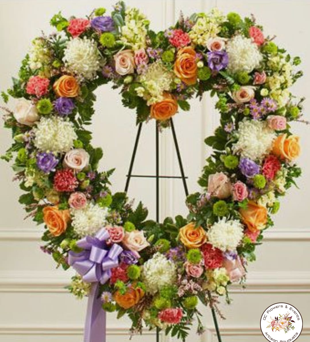 Heart-shaped standing wreath of mixed roses, carnations, and mums in soft colors with a lavender bow