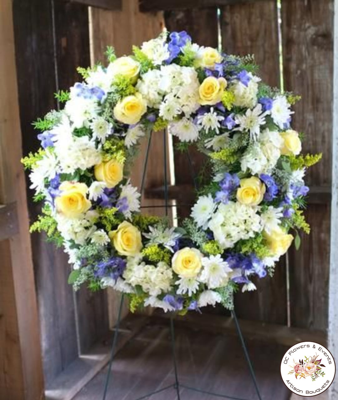 Standing wreath of yellow roses, white daisies, and purple flowers on a metal easel