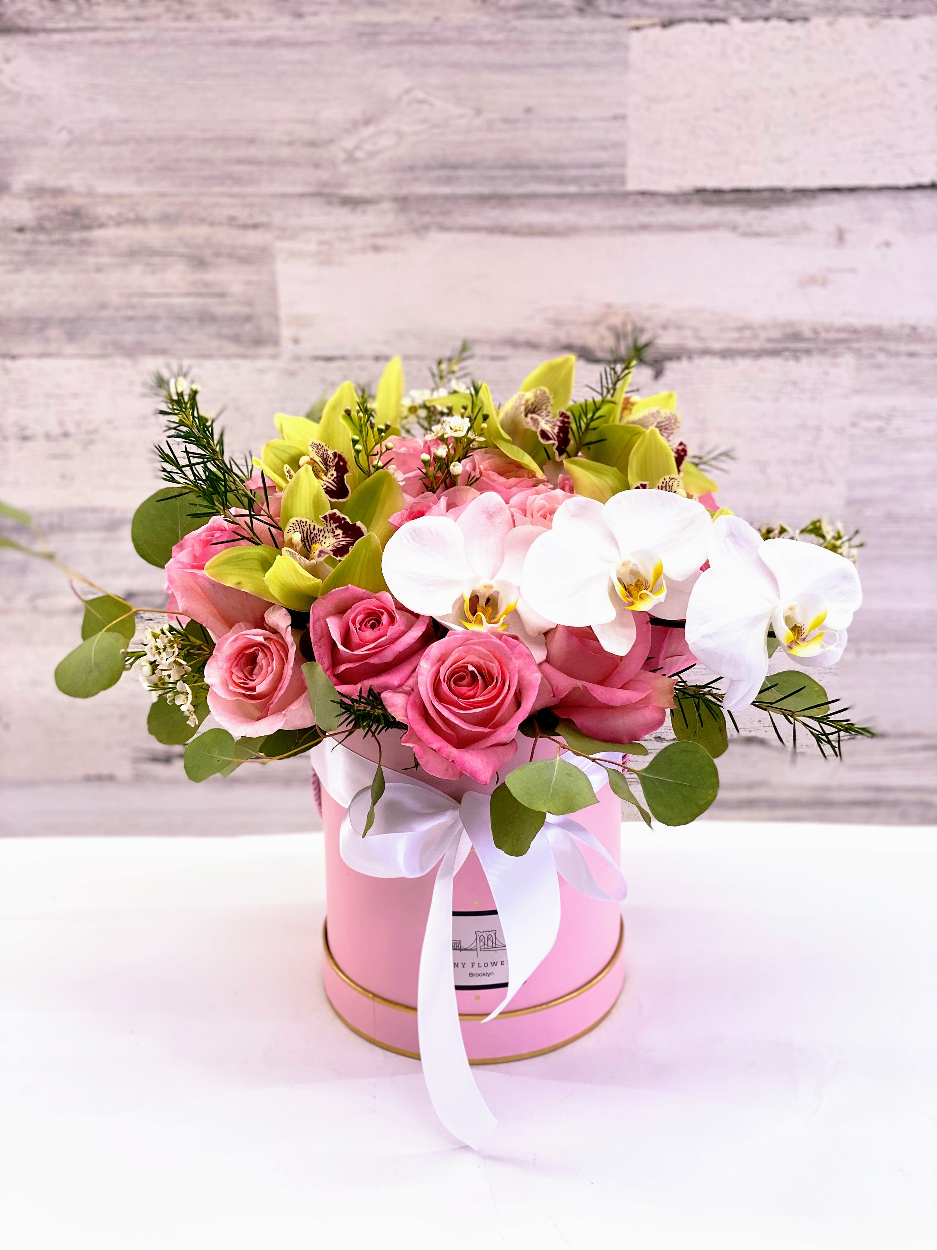 Pink roses and white orchids arranged in a pink hat box with a ribbon