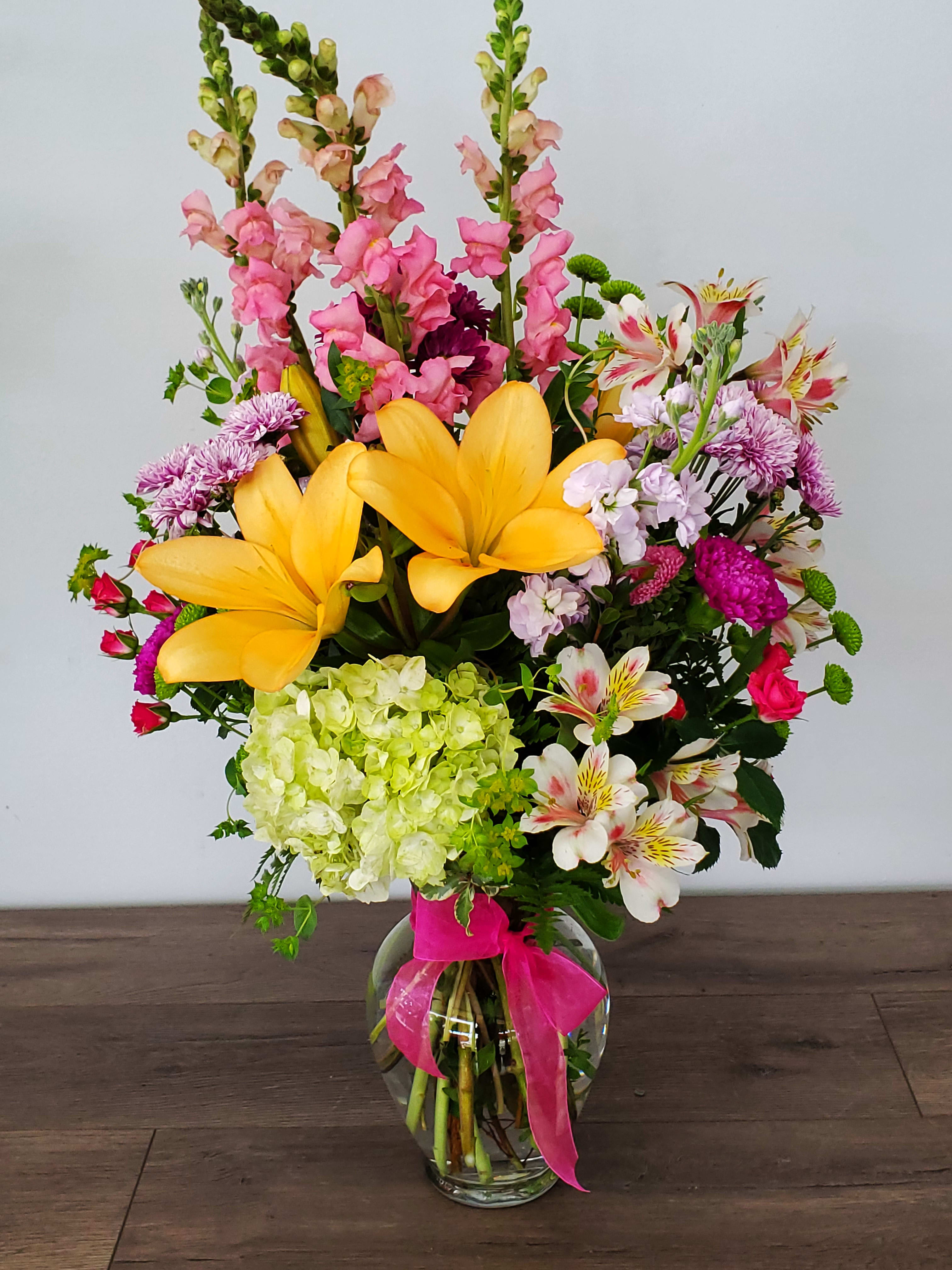 Mixed bouquet with yellow lilies, pink snapdragons, and a glass vase tied with a pink ribbon