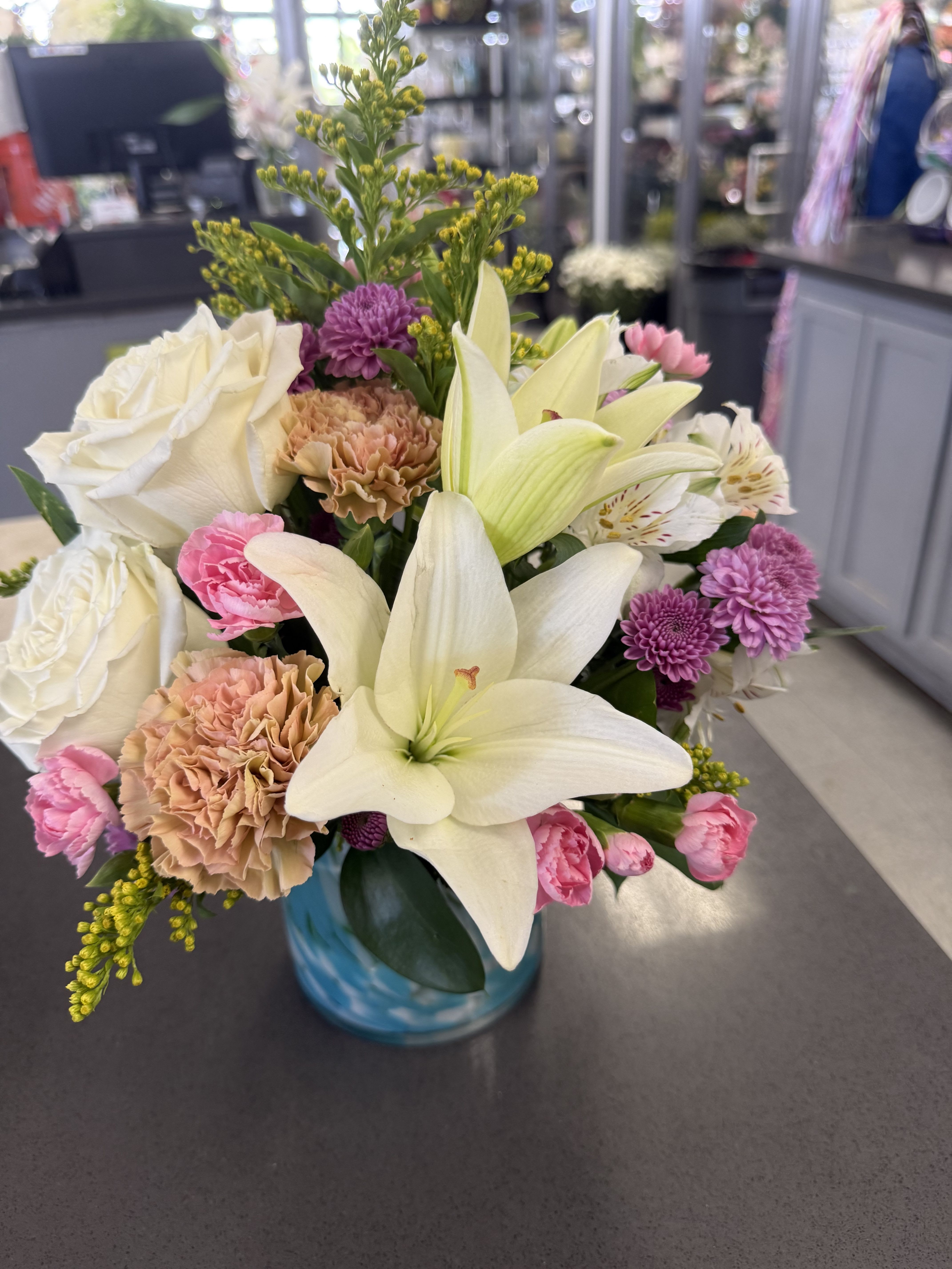 Charm her - Soft pastels meet a modern blue glass vase in this charming flower arrangement-perfect as a birthday bouquet, thank-you gift, or spring celebration arrangement. The mix of large white lilies and creamy roses with blush pink carnations and lavender-purple chrysanthemums creates a gentle, romantic impression that will make her feel truly seen and celebrated.  This rounded arrangement also includes white alstroemeria, pale yellow accents, and bright yellow filler flowers that add height and an airy, fresh-cut texture above the fuller blooms. Arranged in a cool blue glass vase (available in clear glass as noted), the palette of white, pink, purple, yellow, and a hint of blue reads soft yet cheerful and versatile for everyday gifting.  Ideal for the person who appreciates a lightly romantic, upbeat bouquet, this design arrives ready to display and brings a burst of springtime color to any room. Care: keep water fresh and trim stems every few days to extend vase life.