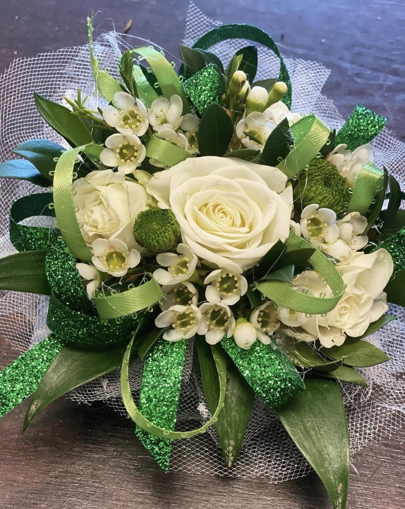 Elegant Verdant Rose - A timeless corsage featuring a pristine white rose nestled among fresh, lush greenery, creating a look that is both classic and naturally beautiful. Accented with flowing silver ribbons and shimmering sparkling green ribbon details, this piece catches the light with every movement, adding a touch of glamour to its soft, organic design. The combination of crisp white, rich green, and metallic shimmer makes this corsage effortlessly elegant—perfect for proms, weddings, or any special occasion where a refined, graceful accessory is desired.