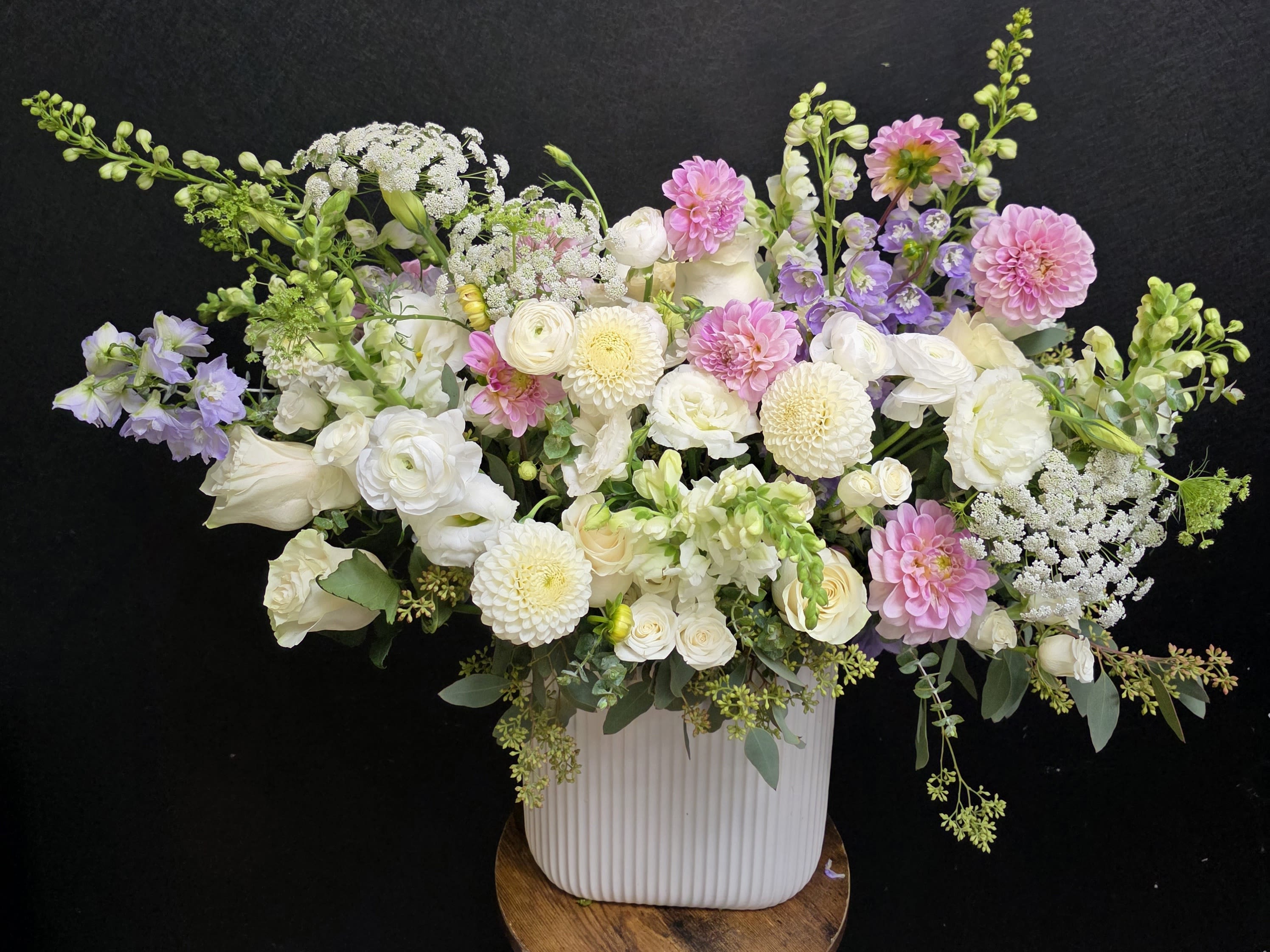 Blushing Meadow - Soft blush roses and lavender delphinium rise from a cloud of white blooms in this garden-style flower arrangement-perfect as a wedding centerpiece or a springtime birthday bouquet. The large, airy composition in a white ribbed vase reads romantic and gently whimsical; the person you're celebrating will feel seen by the soft pastels and the graceful, lightly asymmetrical silhouette.  The arrangement layers rounded roses, ranunculus, and dahlias with taller snapdragons and delphinium spires, finished with delicate branching accents and cheerful daisies for texture. Greens peek through to brighten the palette of white, pink, and purple; the vase sits on a simple wooden stand to elevate the display and give it presence on a table or entry console.  Ideal for weddings, anniversaries, spring celebrations, or as a thoughtful everyday gift, this piece makes a confident statement without overwhelming the room. Designed at a large scale to work as a centerpiece, it arrives ready to display-keep fresh water in the vase and trim stems a half-inch every few days for longest enjoyment.