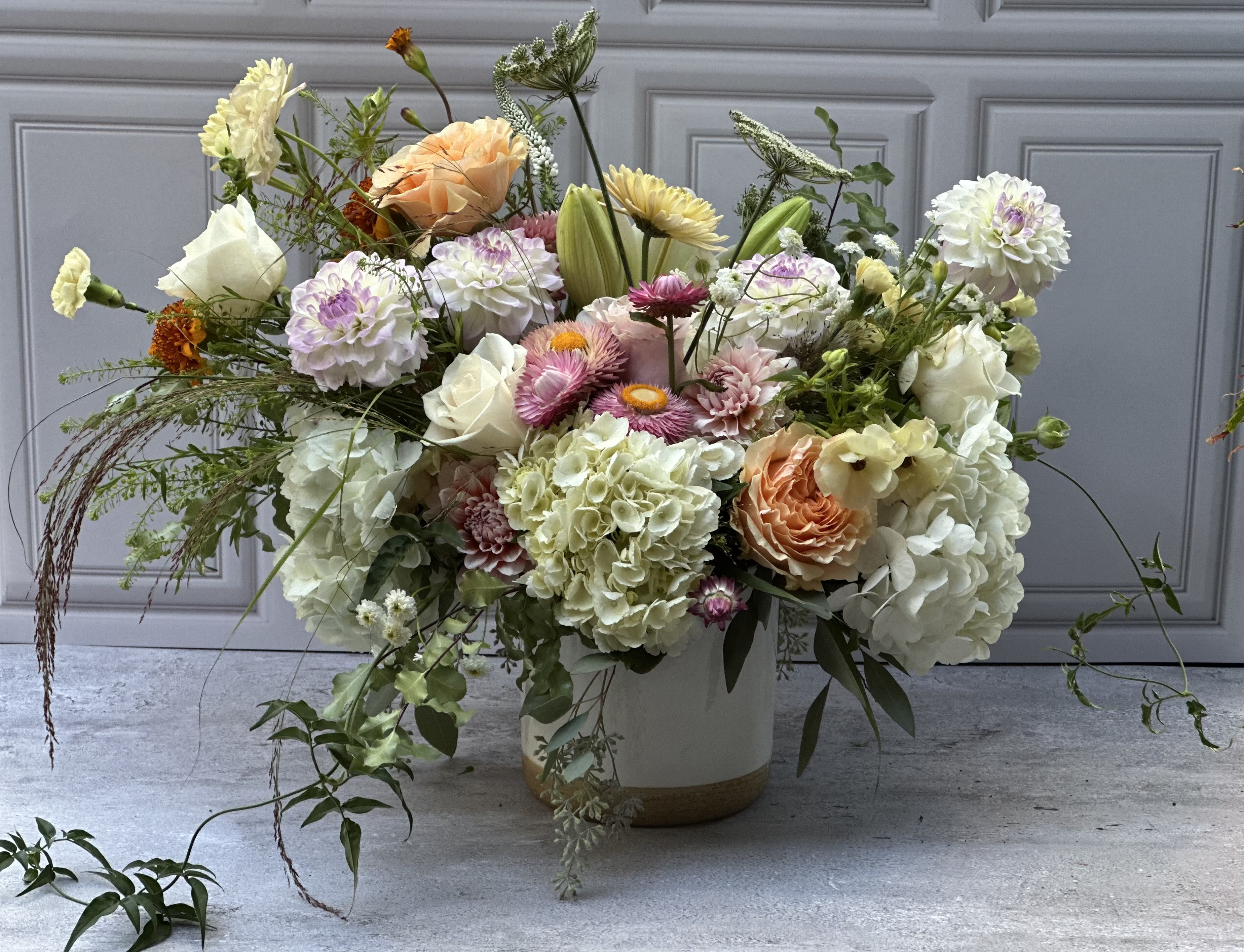 The Grand - A generous, garden-style flower arrangement built in a white ceramic vase, perfect as a large birthday bouquet, anniversary flowers, or a springtime celebration for someone special. The Grand greets the recipient with a soft, romantic palette of cream, blush, peach and soft pink-the kind of arrangement that makes someone feel celebrated and truly seen the moment it's set down.  Rounded hydrangea clusters and classic roses anchor the composition while dahlias, lilies, chrysanthemums and cheerful daisies add layered texture and movement; taller stems and delicate filler create an airy, loose silhouette rather than a tightly structured dome. The white ceramic vase provides a clean, elegant base that suits both casual rooms and more formal tabletops; the overall scale reads generous and abundant without feeling crowded.  Ideal for marking birthdays, anniversaries, or simply saying "I'm thinking of you," this arrangement arrives ready to display. For best longevity, refresh the water every two days and trim stems slightly when you change the water to keep blooms looking their freshest.