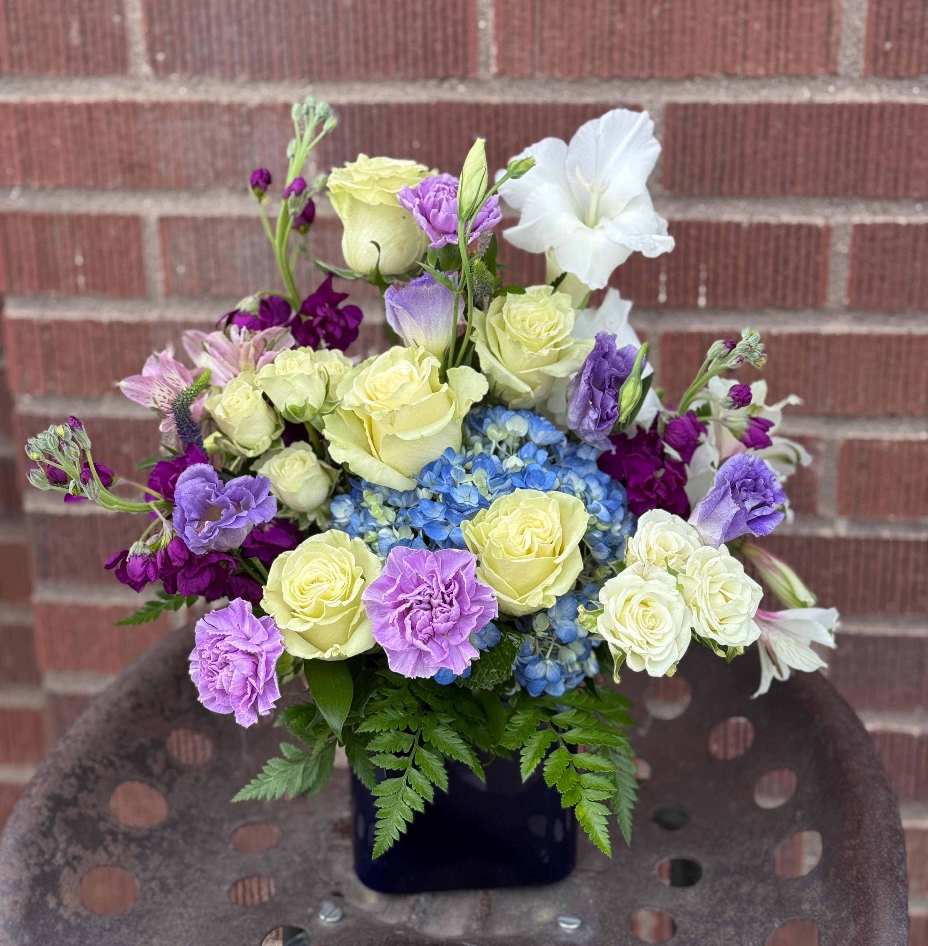 Bouquet of yellow roses, blue hydrangea, and purple flowers in a dark vase