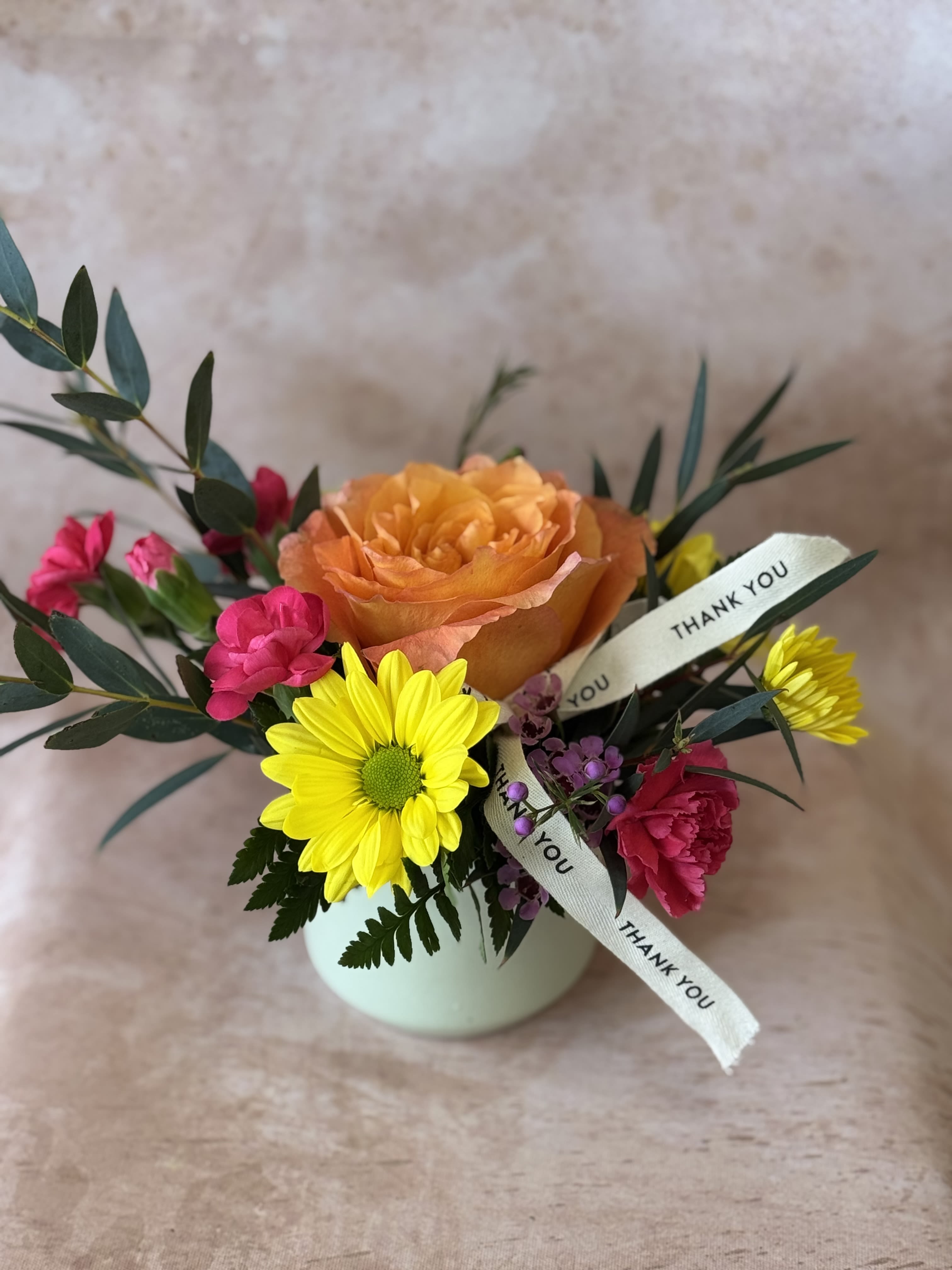 Thank you very much! - A large orange rose anchors this compact "Thank you" flower arrangement, making an immediate, cheerful statement for a thank-you gift or Admin Professionals' Week surprise. Bright yellow daisy-style blooms and vivid pink flowers surround the rose for lively contrast, while small purple filler blossoms add texture and a playful, hand-picked feel-the kind of bouquet that makes the person you're thanking feel genuinely seen.  Set in a soft round ceramic vase and finished with a cream printed fabric ribbon that reads THANK YOU, this friendly arrangement reads as both professional and personal. Colors include orange, yellow, pink, and white; flowers include daisies, carnations, roses, and assorted seasonal fillers. Its compact size suits desks, reception areas, or a kitchen table-an easy-to-place token of appreciation that looks fresh with minimal care.