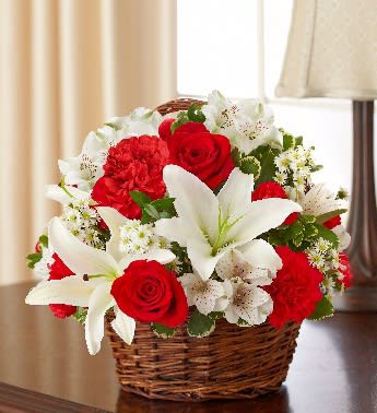 Red roses, red carnations, and white lilies arranged in a wicker basket on a table