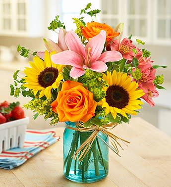 Bright bouquet of sunflowers, orange roses, and pink lilies in a blue mason jar vase with raffia bow
