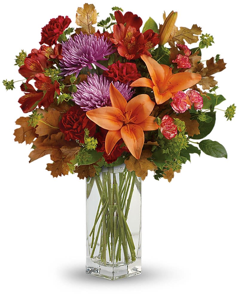 Autumn arrangement of orange lilies, purple mums, and red flowers with brown leaves in a tall glass vase