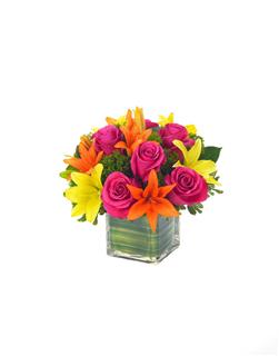 Bright pink roses with orange and yellow lilies in a cube glass vase with green leaf wrap.