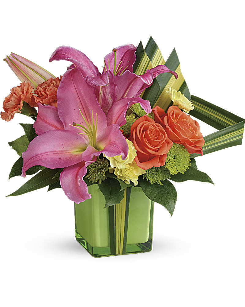 Bright arrangement of pink lilies, orange roses and carnations in a green cube vase with striped leaves.