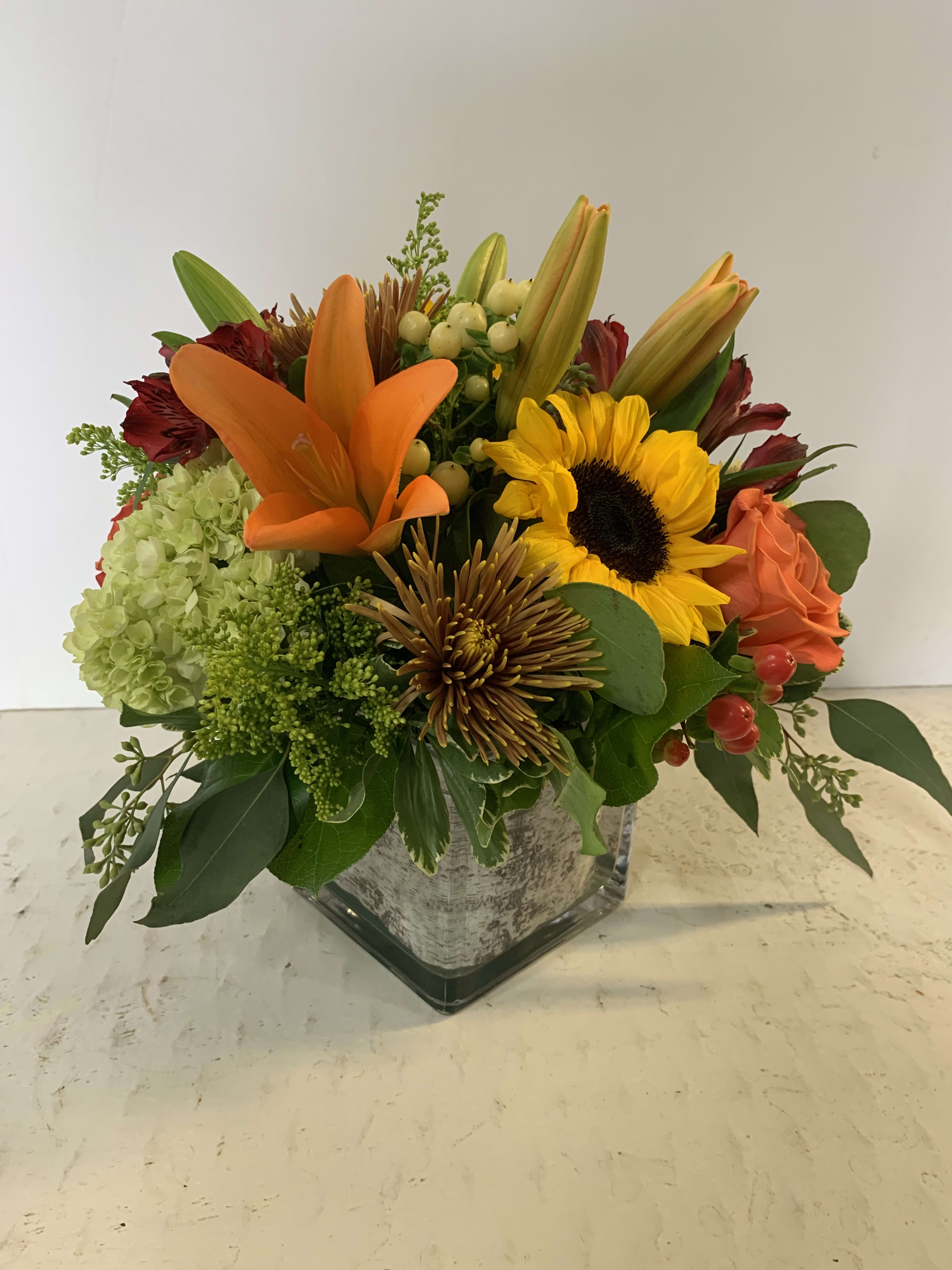 Mixed bouquet with orange lilies, a sunflower, and roses in a square glass vase