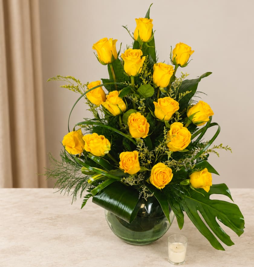 Yellow roses arranged in a glass vase with tropical greenery