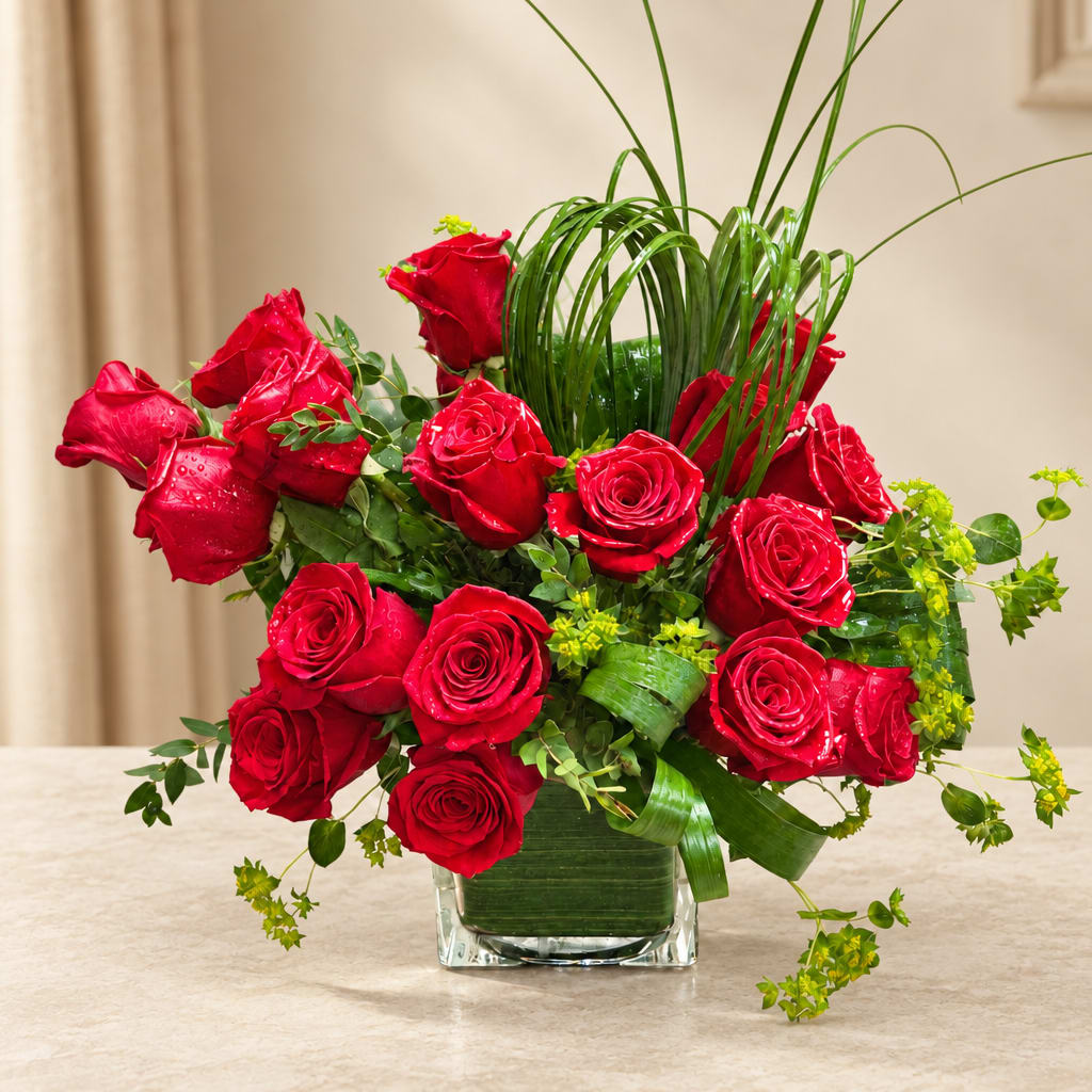 Red roses arranged in a clear square vase with tall grass accents