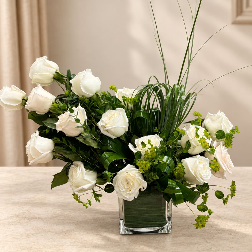White roses arranged in a clear square vase with tall grass accents