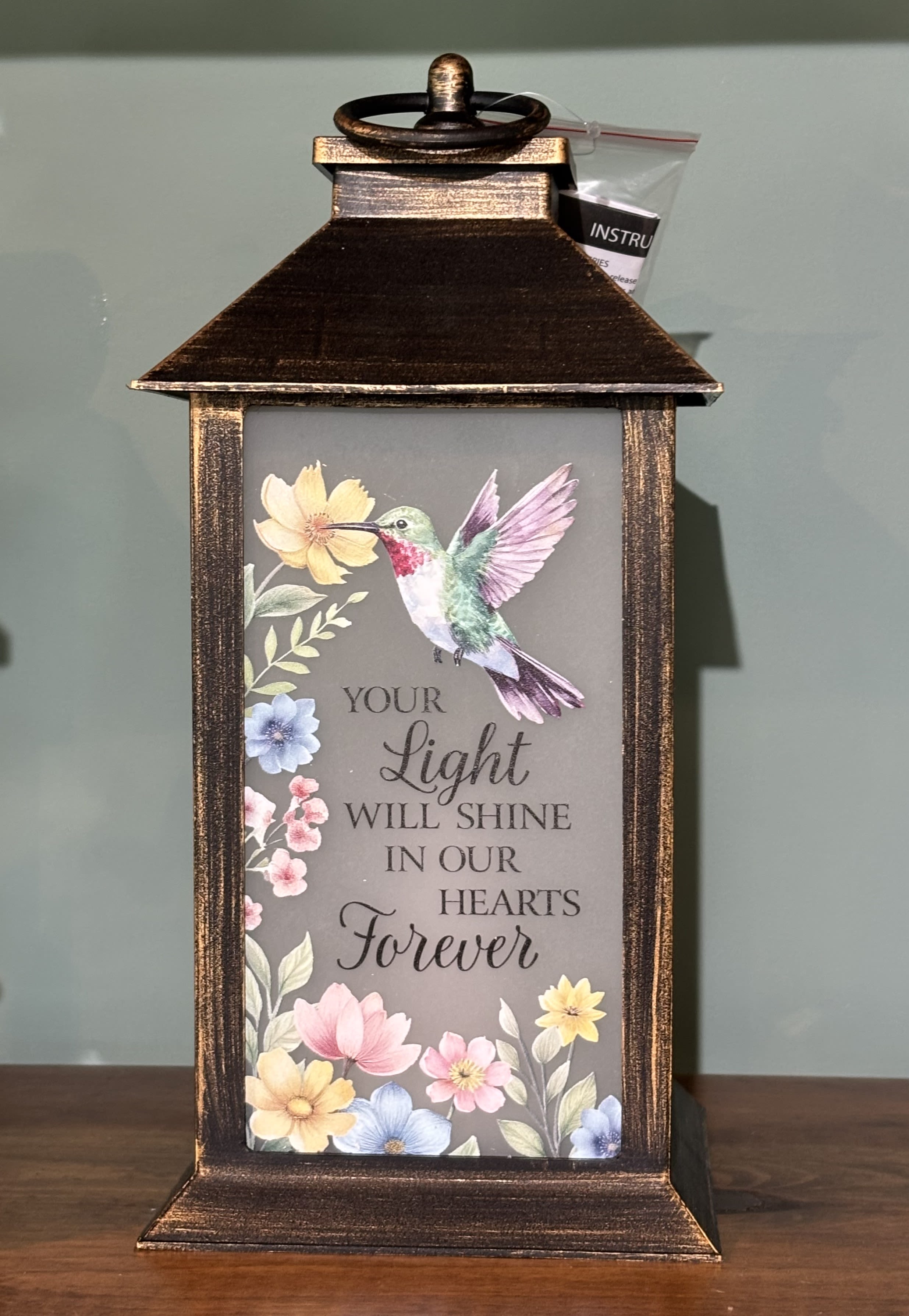 Your Light Will Shine Sympathy Lantern - This beautiful coppery hued high quality polyresin finish lantern surrounds an interior  a realistic LED flickering flame. The lantern has a "frost" look with a lovely pastel florals and hummingbird pictured on the front panel as well as the touching sentiment "Your light will shine in our hearts forever". Lantern is 13"H x 5 1/2"W x 5 1/2" D. A built in automatic timer and 3 AAA batteries are included. This item is suitable for sympathy gifting either at the vistation or the home This item is weather friendly.