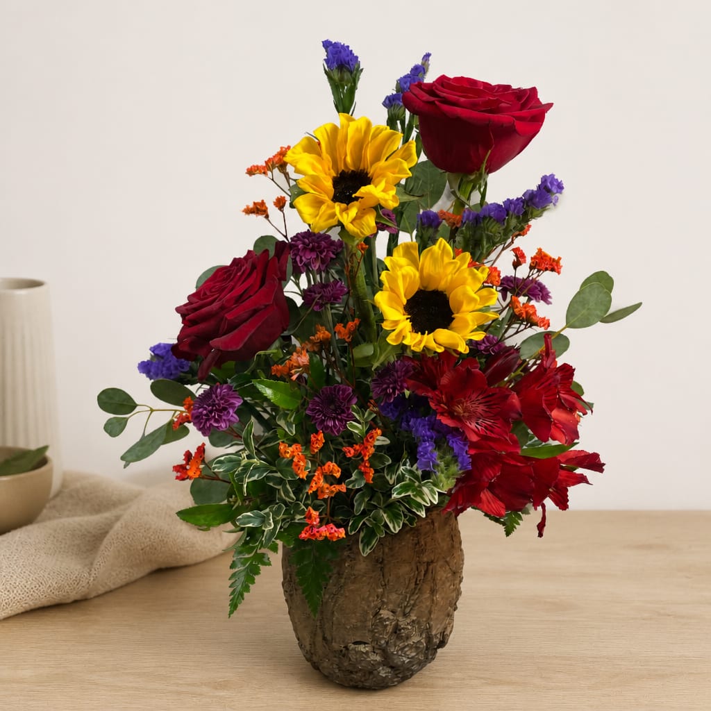 Mixed bouquet with red roses, yellow sunflowers, and purple accents in a rustic vase