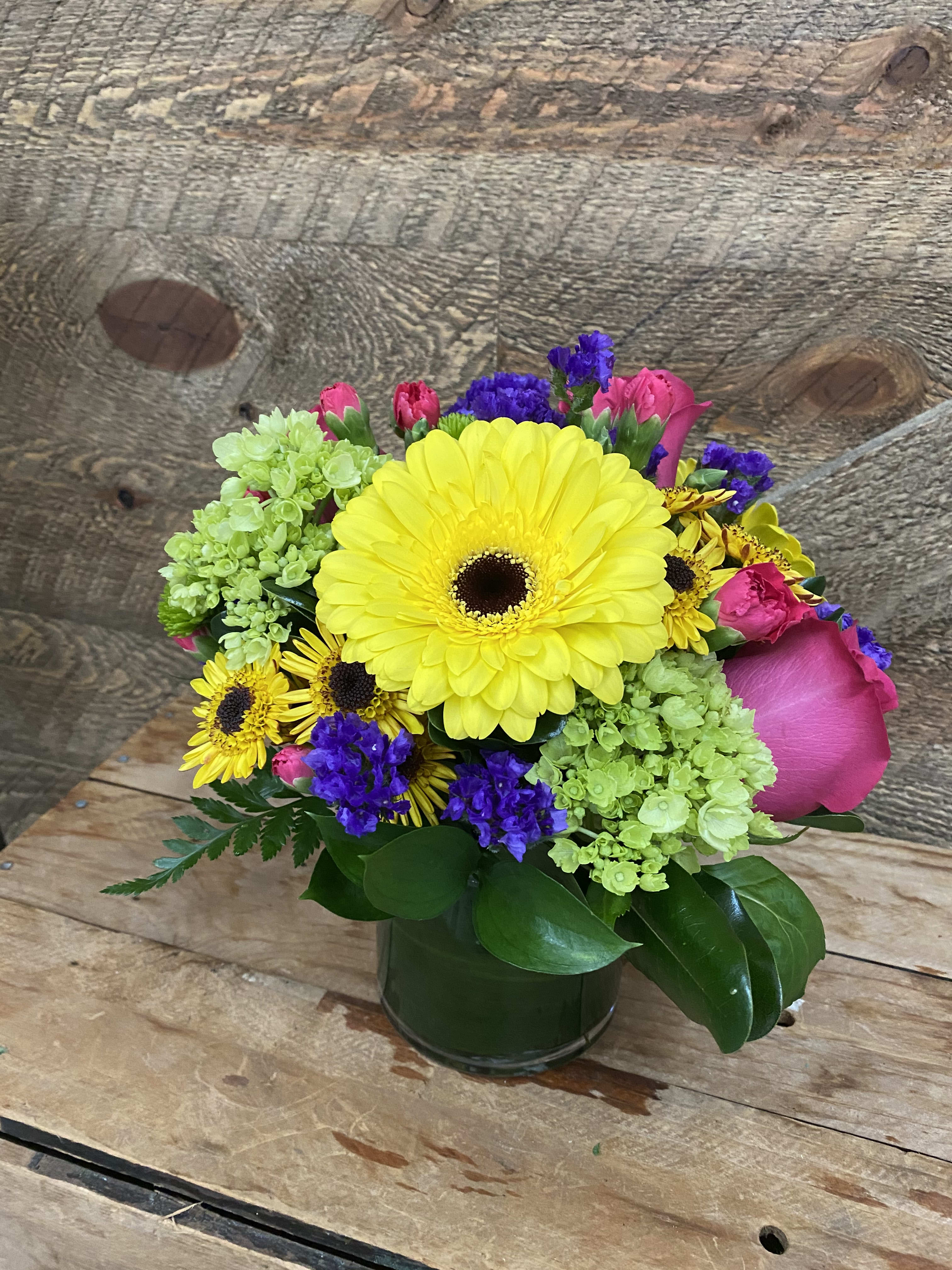 Mixed bouquet with yellow gerbera daisies, pink roses, and purple accents in a glass vase