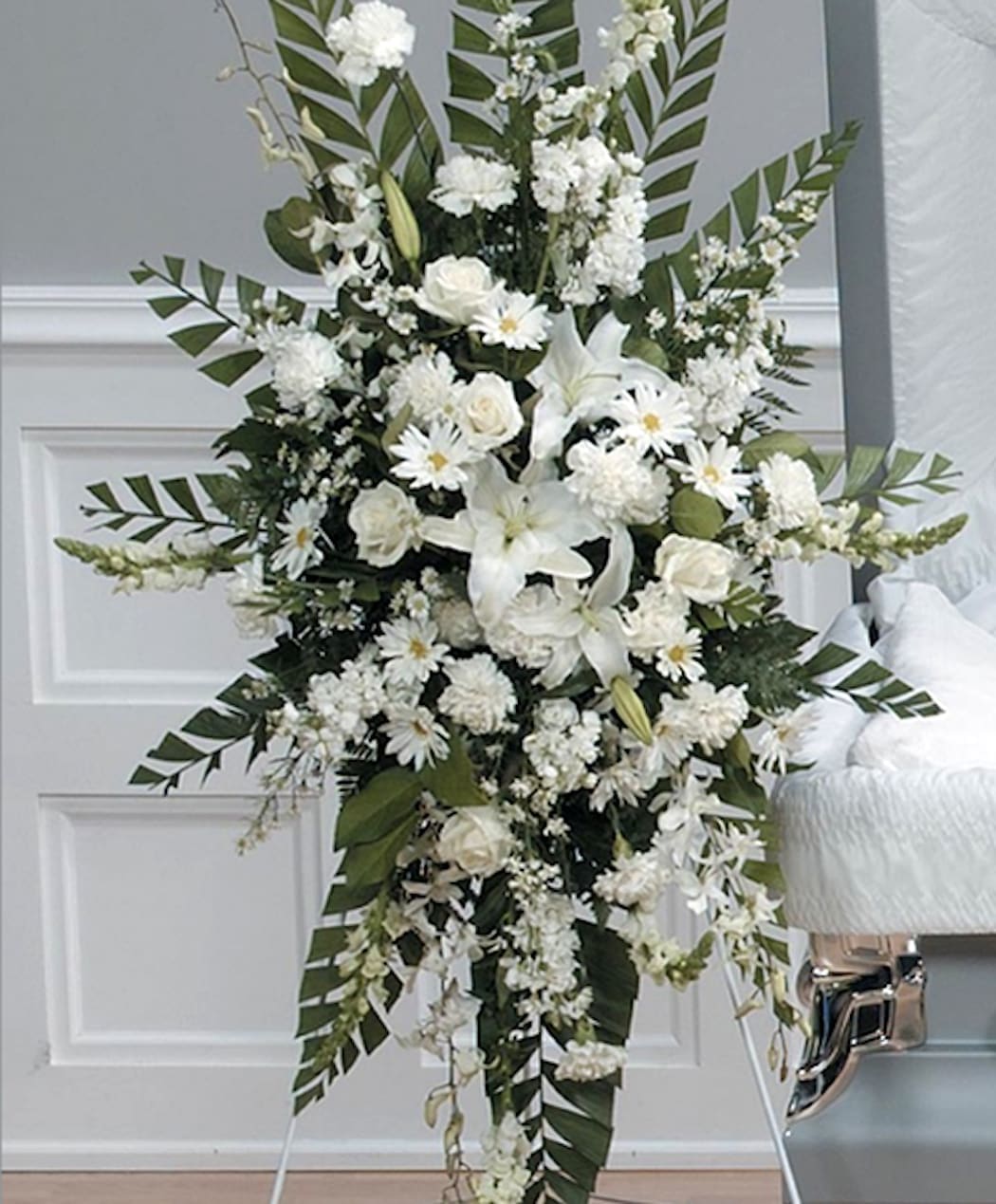 Tall all-white standing spray of lilies, roses, carnations, and daisies on an easel beside a casket