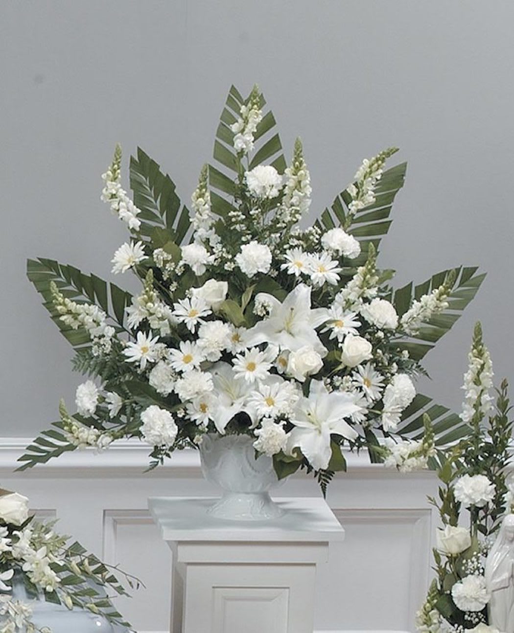 Tall white arrangement with lilies, roses, carnations, and daisies in a white urn with palm leaves