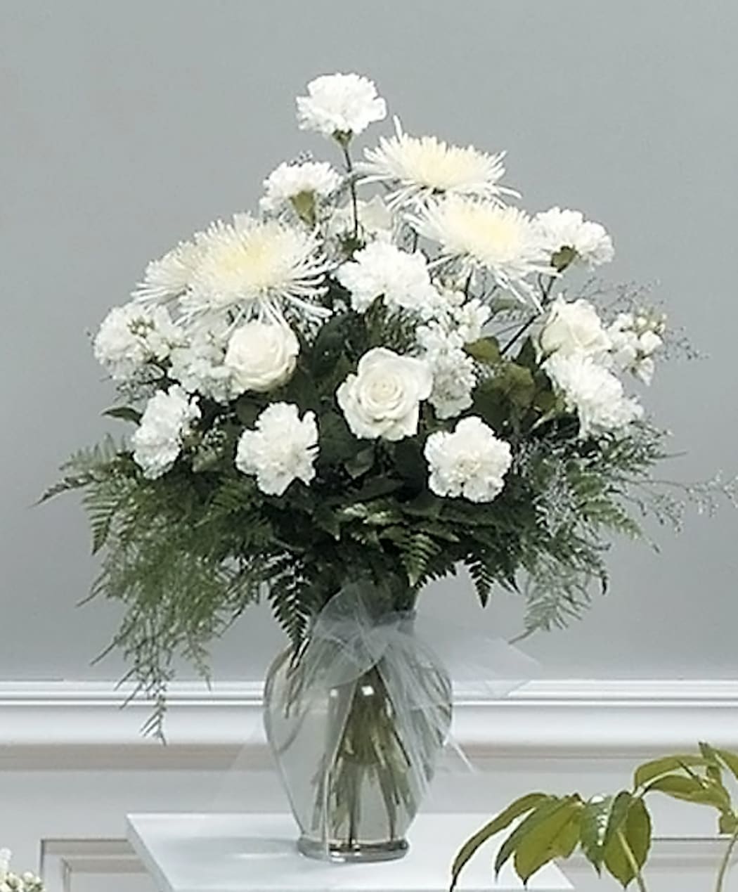 Tall arrangement of white roses, carnations, and spider mums in a clear glass vase with bow