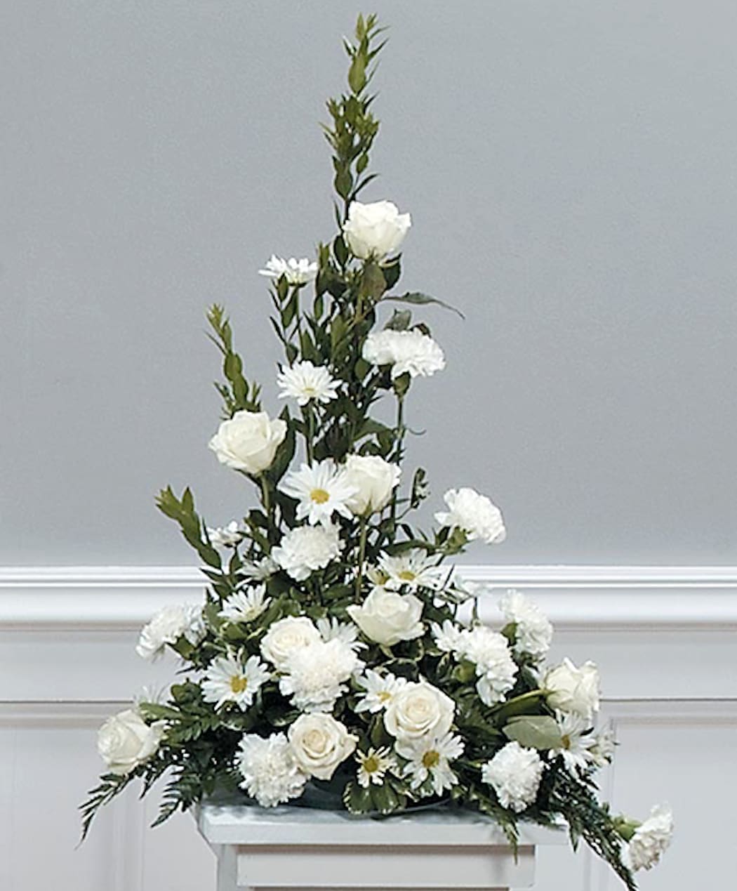 Tall white arrangement of roses, daisies, and carnations in a low container on a pedestal.