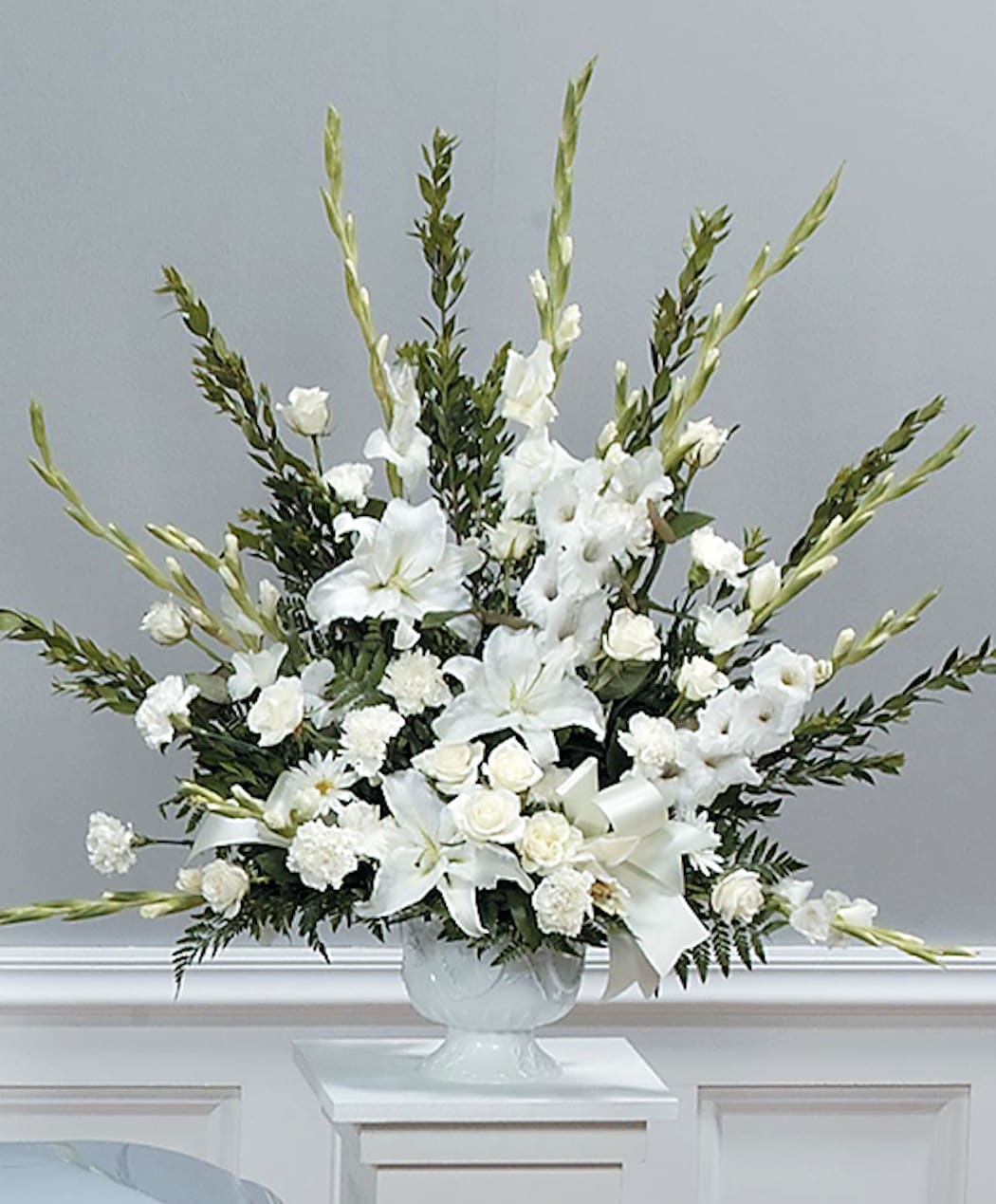 Tall all-white arrangement with lilies, roses, carnations, and gladiolus in a white urn.